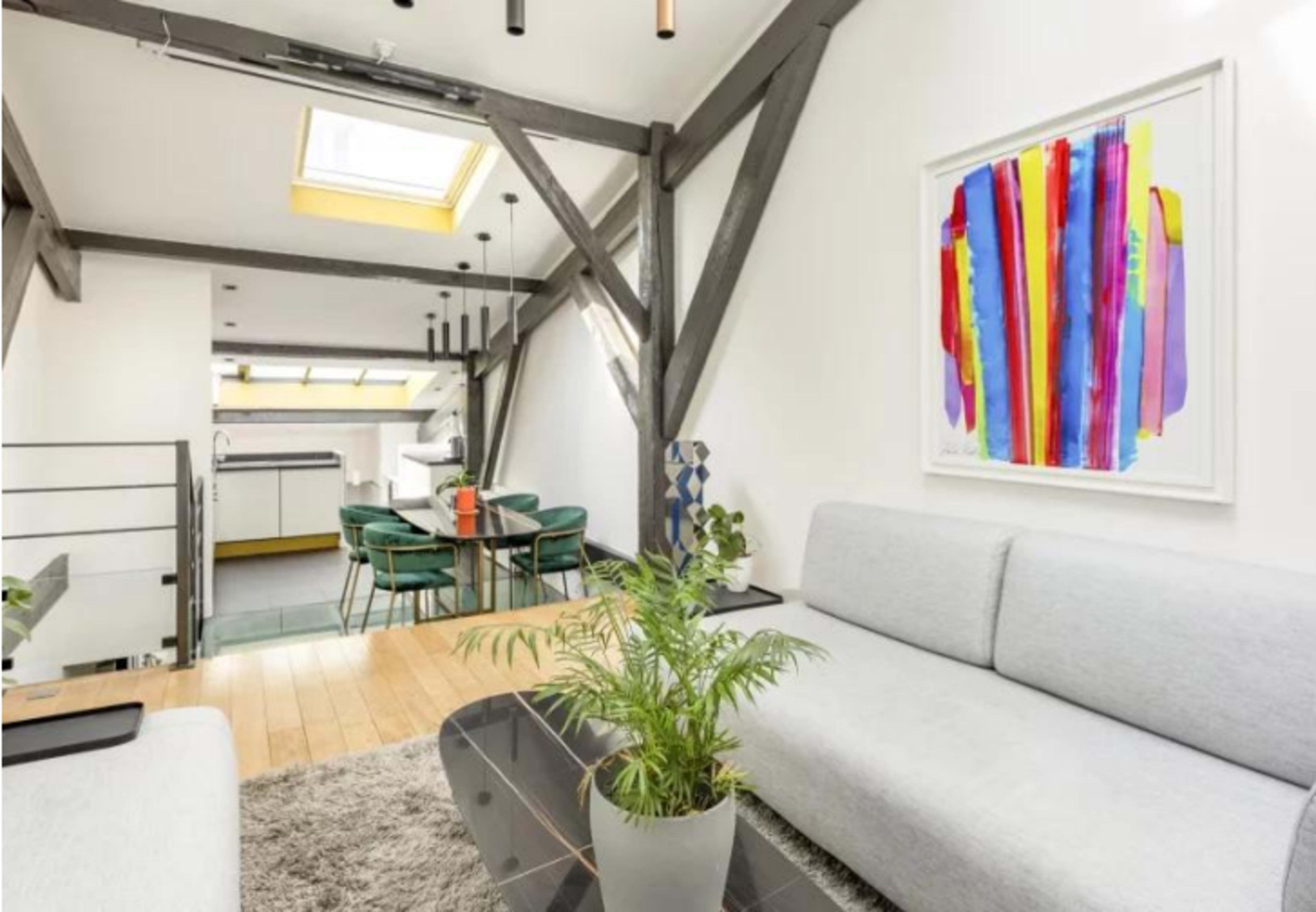The image shows a modern living area with a gray sofa, a glass coffee table, a potted plant, and a colorful piece of art on the wall, leading to a dining area and kitchen in the background.