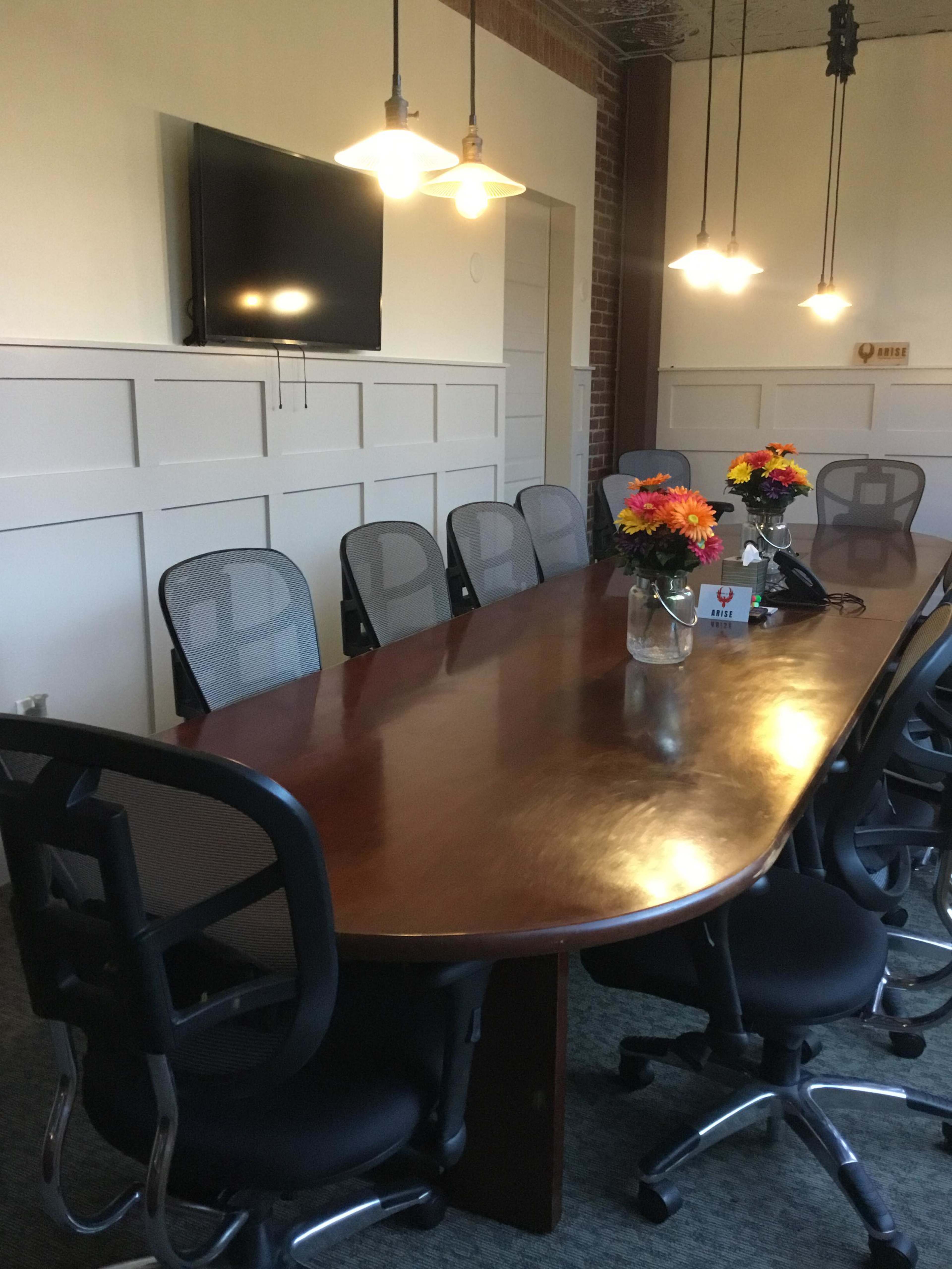 A long wooden conference table is surrounded by black ergonomic chairs in a well-lit room with wall-mounted decor and a television.