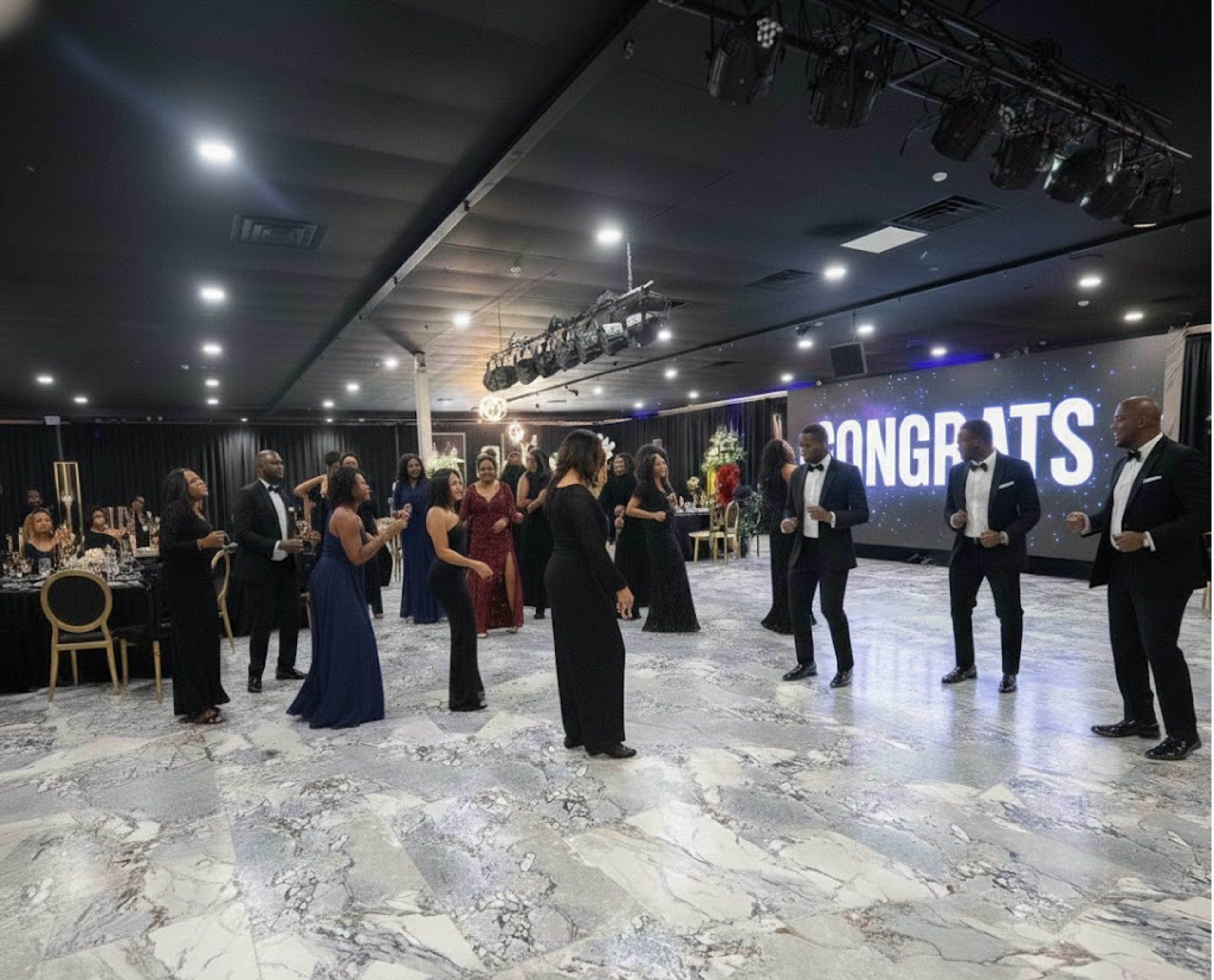 A group of elegantly dressed people are dancing together in a well-lit event space with a "Congrats" backdrop.