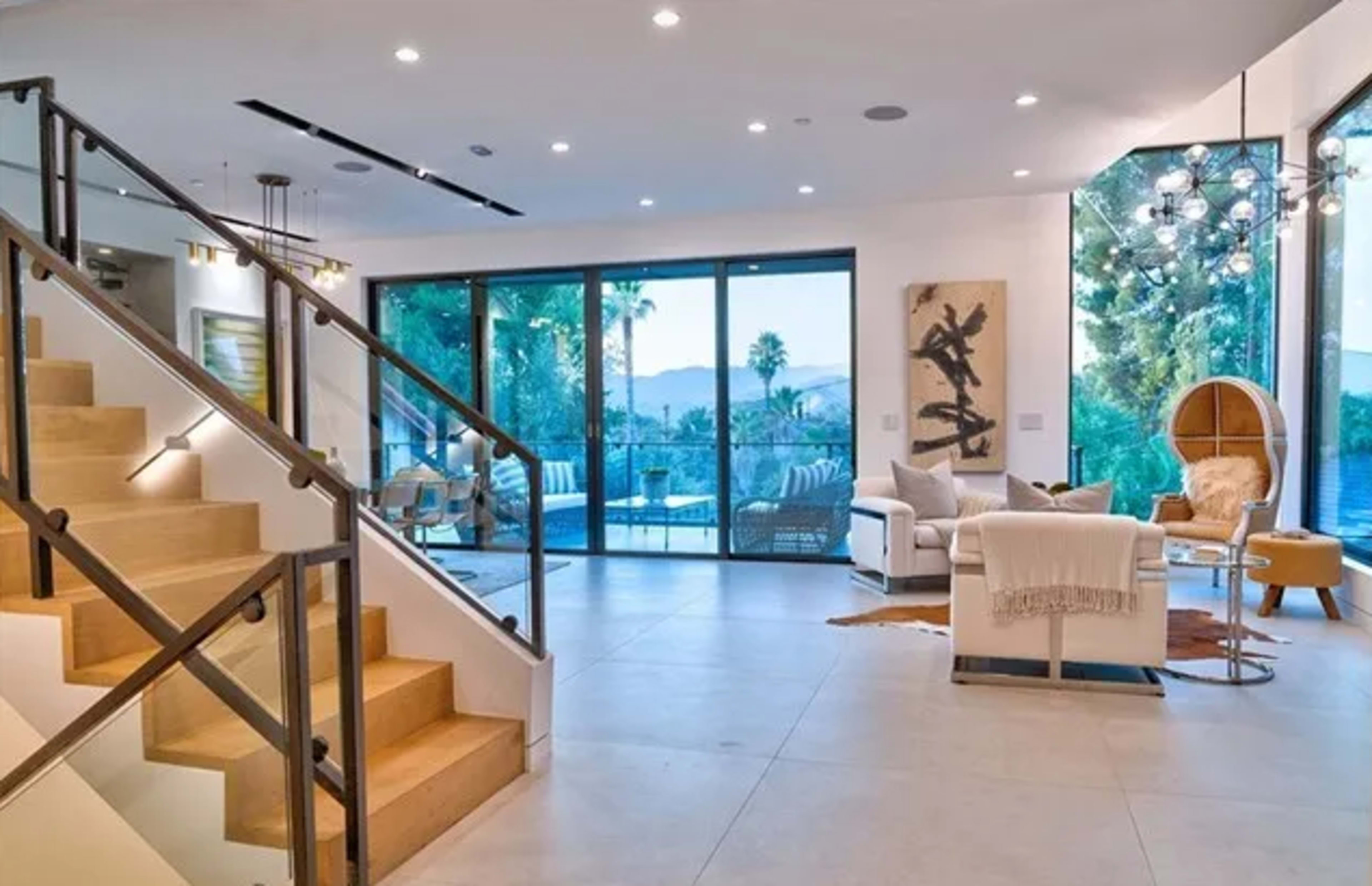 The image shows a modern living room with large glass doors leading to a patio, a staircase with wooden steps, and contemporary furnishings.