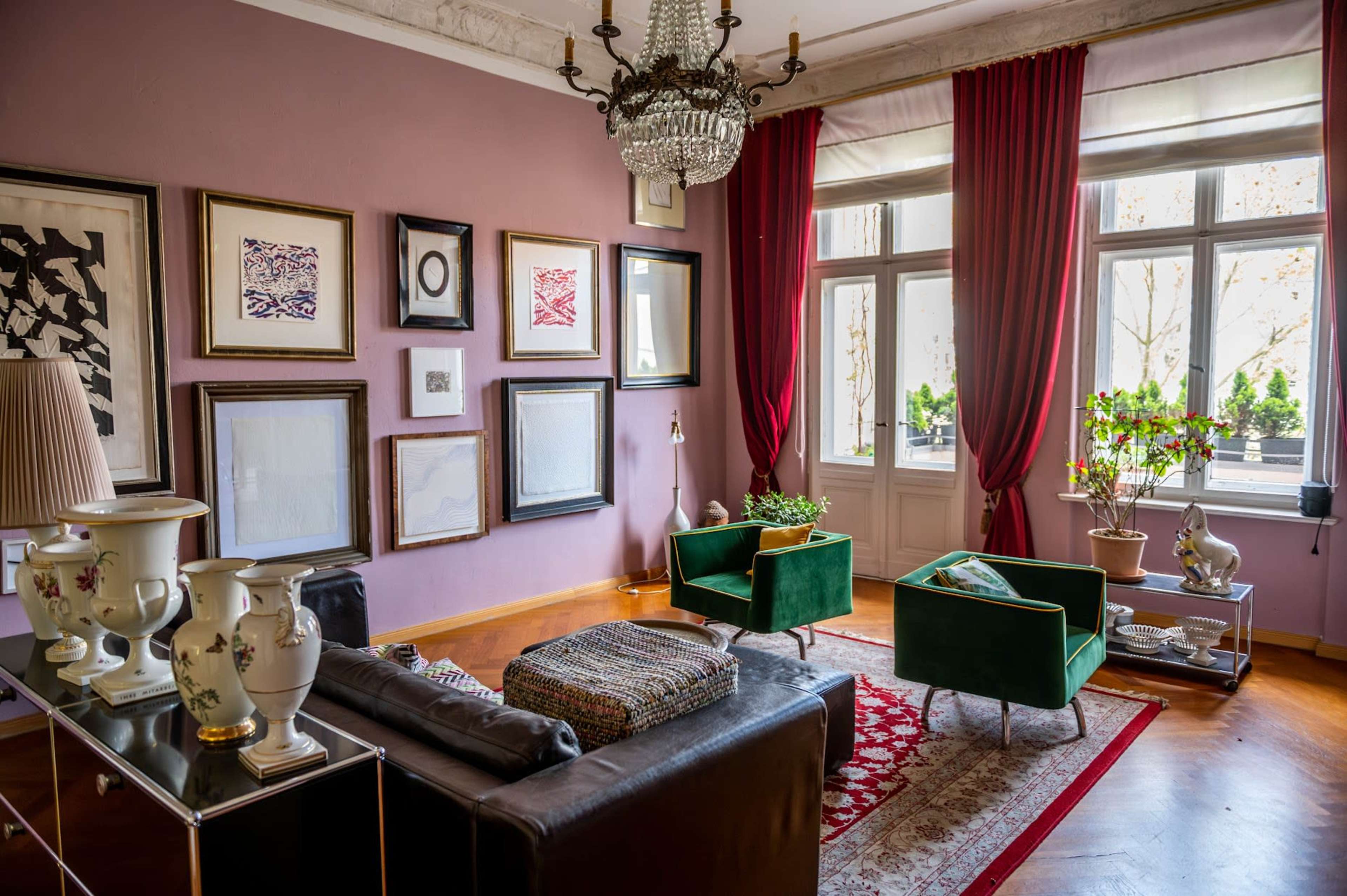 The image shows a well-decorated living room featuring a chandelier, framed artwork on the walls, and two green armchairs beside a window with red curtains.