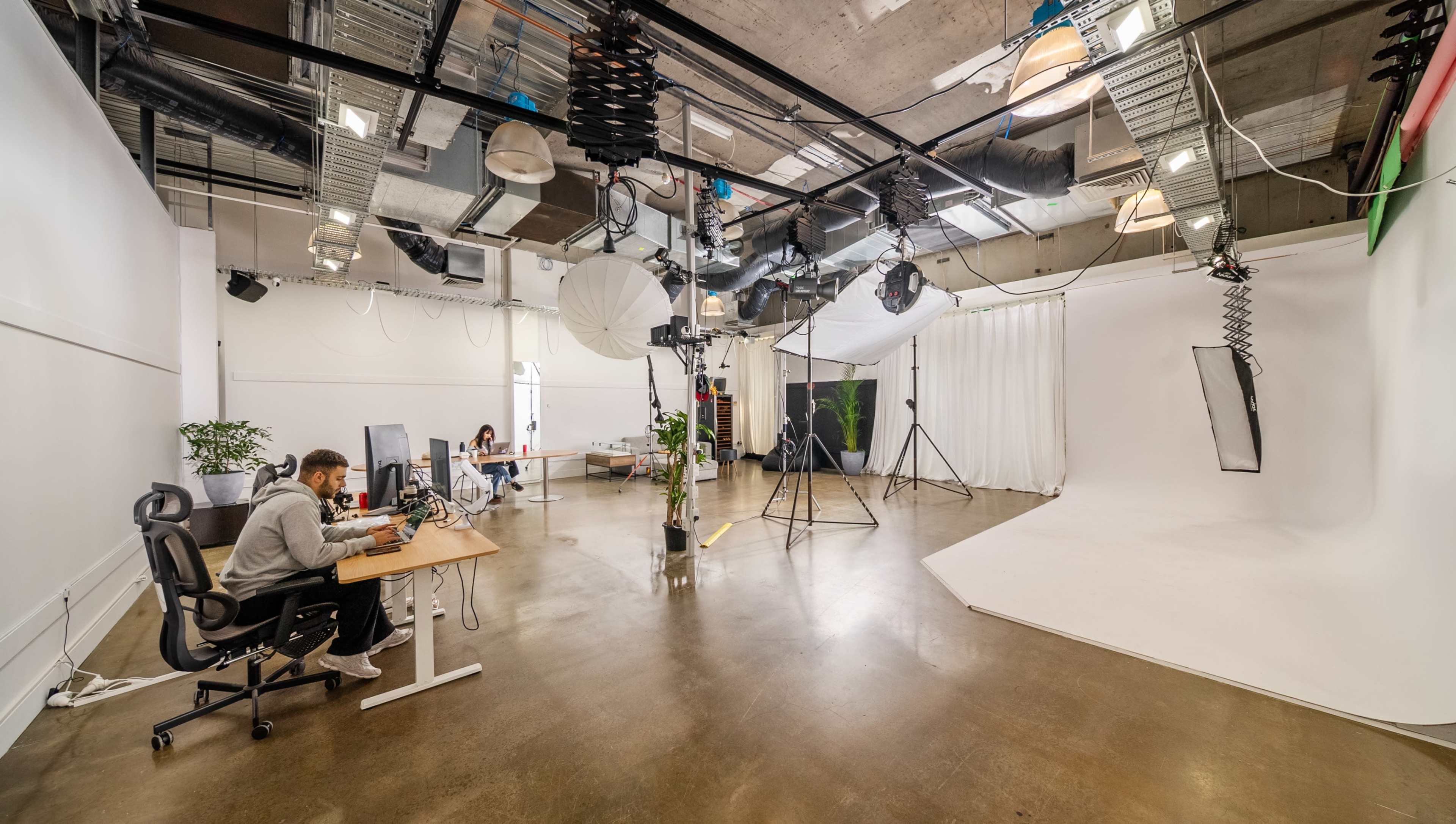 The image shows a spacious studio environment with a white backdrop, lighting equipment, and two individuals working at desks.