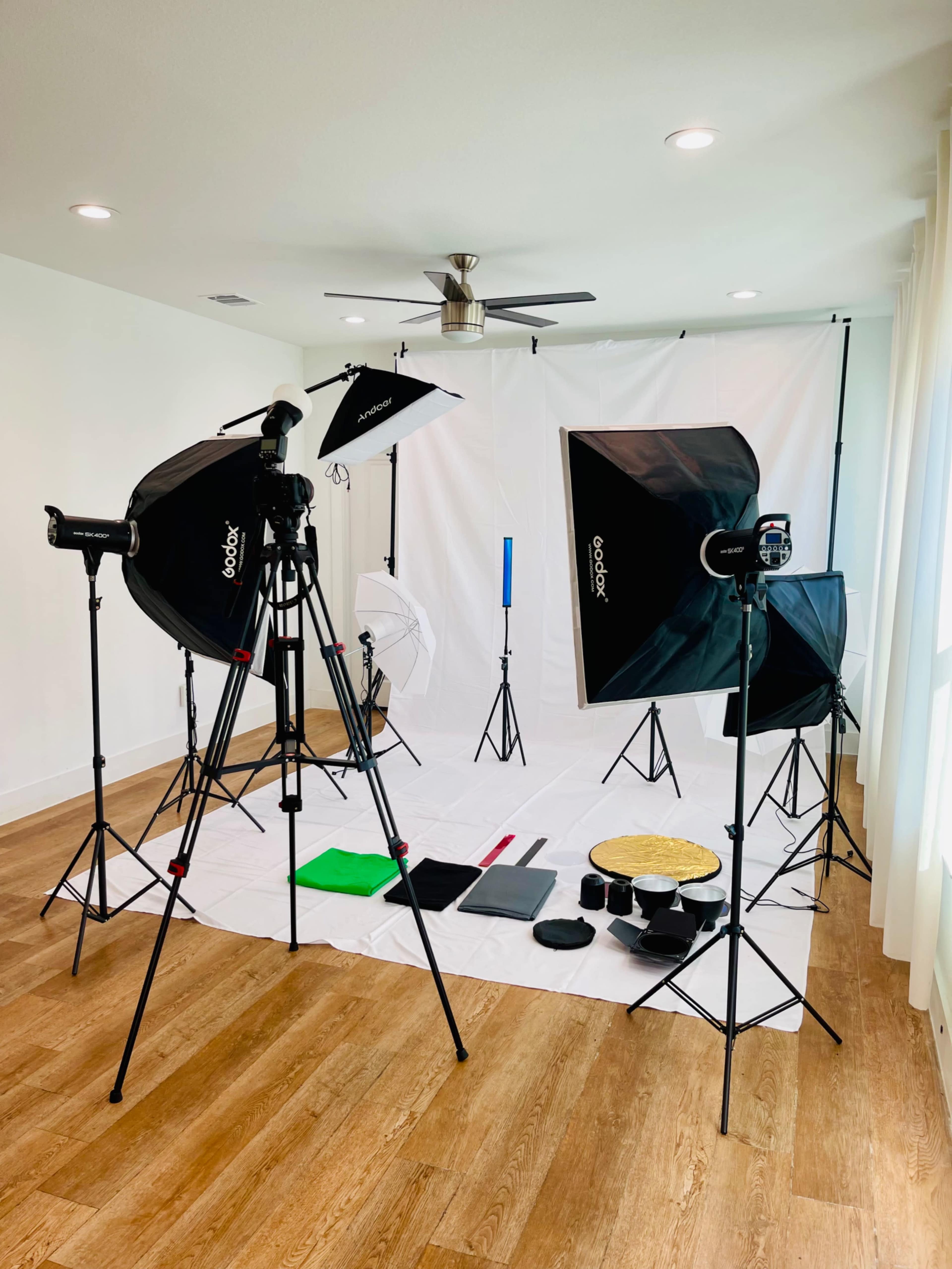 A photography studio setup features softbox lights, tripods, and a white backdrop with various colored props arranged on the floor.