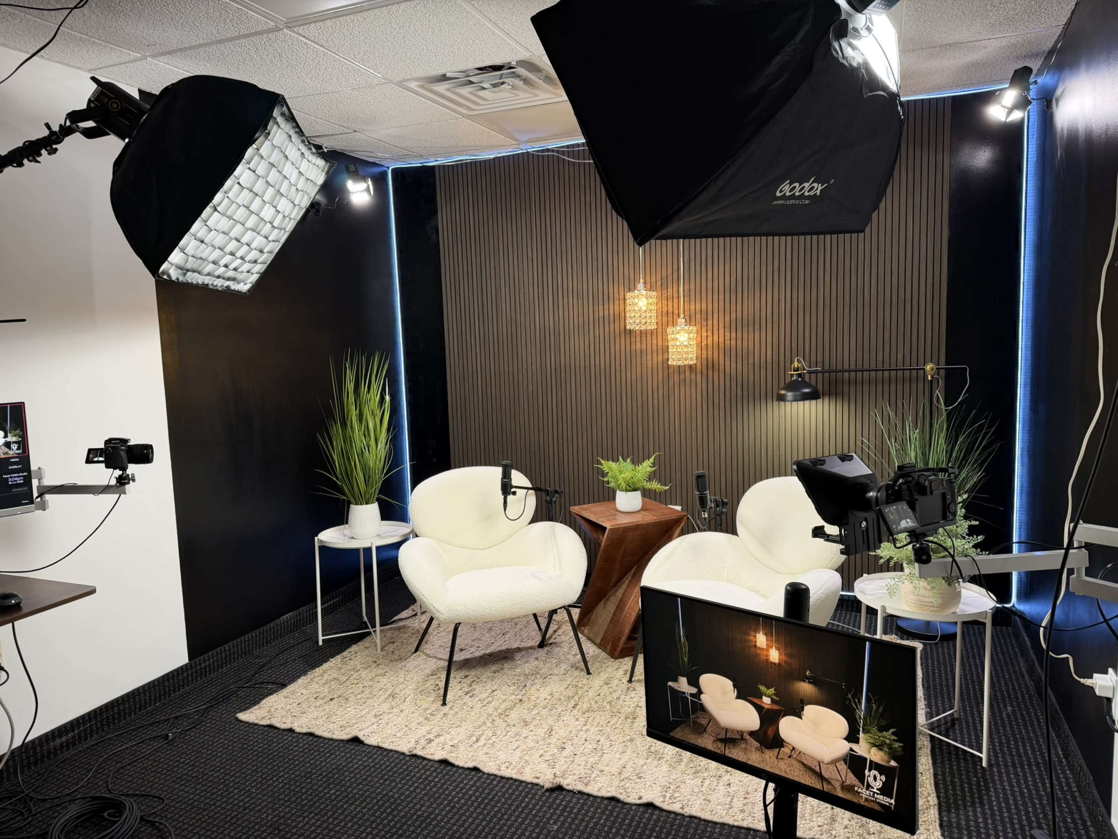 The image shows a studio setup with two white chairs, a small table, plants, and lighting equipment arranged against a dark backdrop.