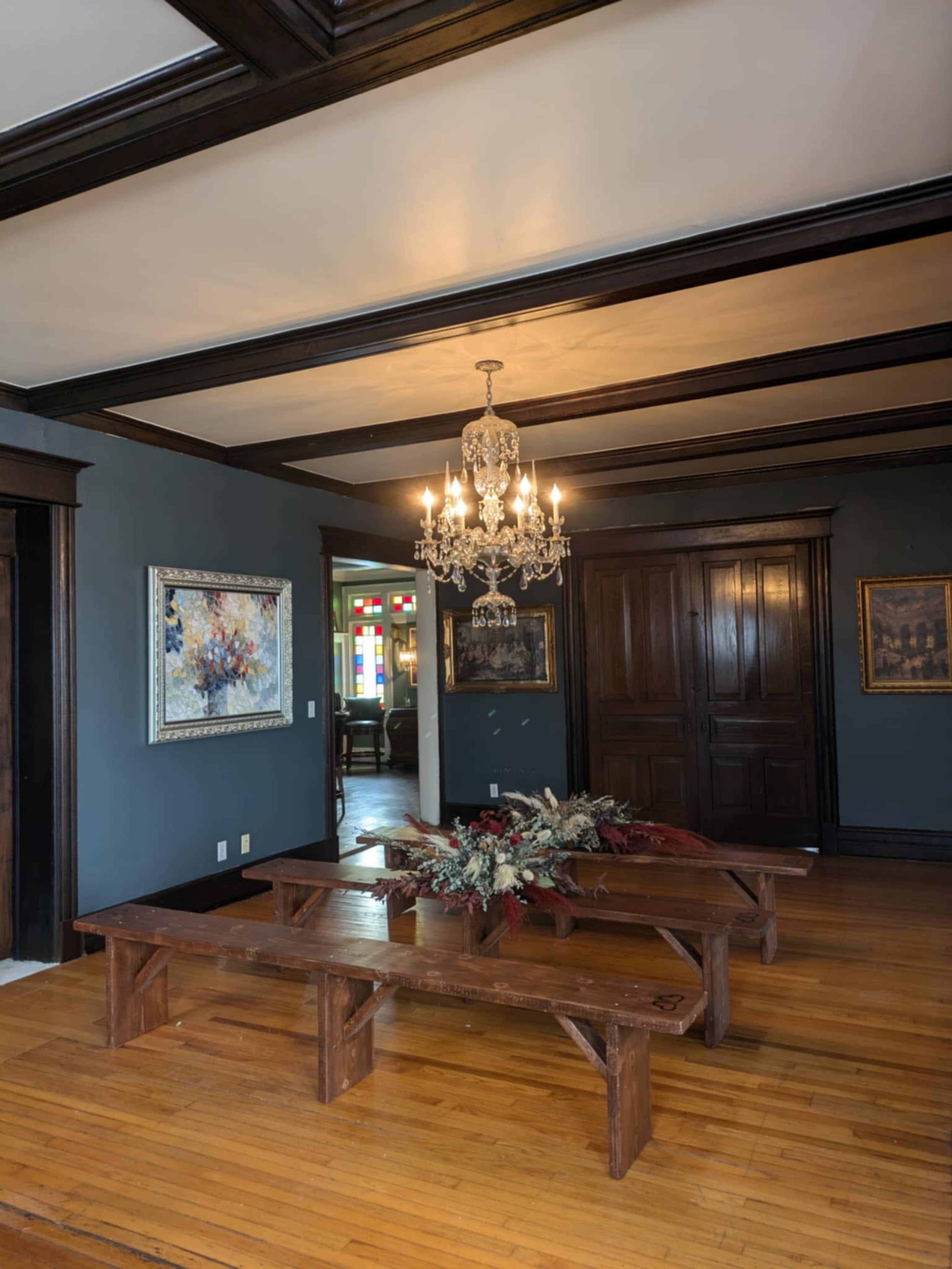 The room features dark wooden beams, a crystal chandelier, a wooden bench table, and framed artworks on the walls.