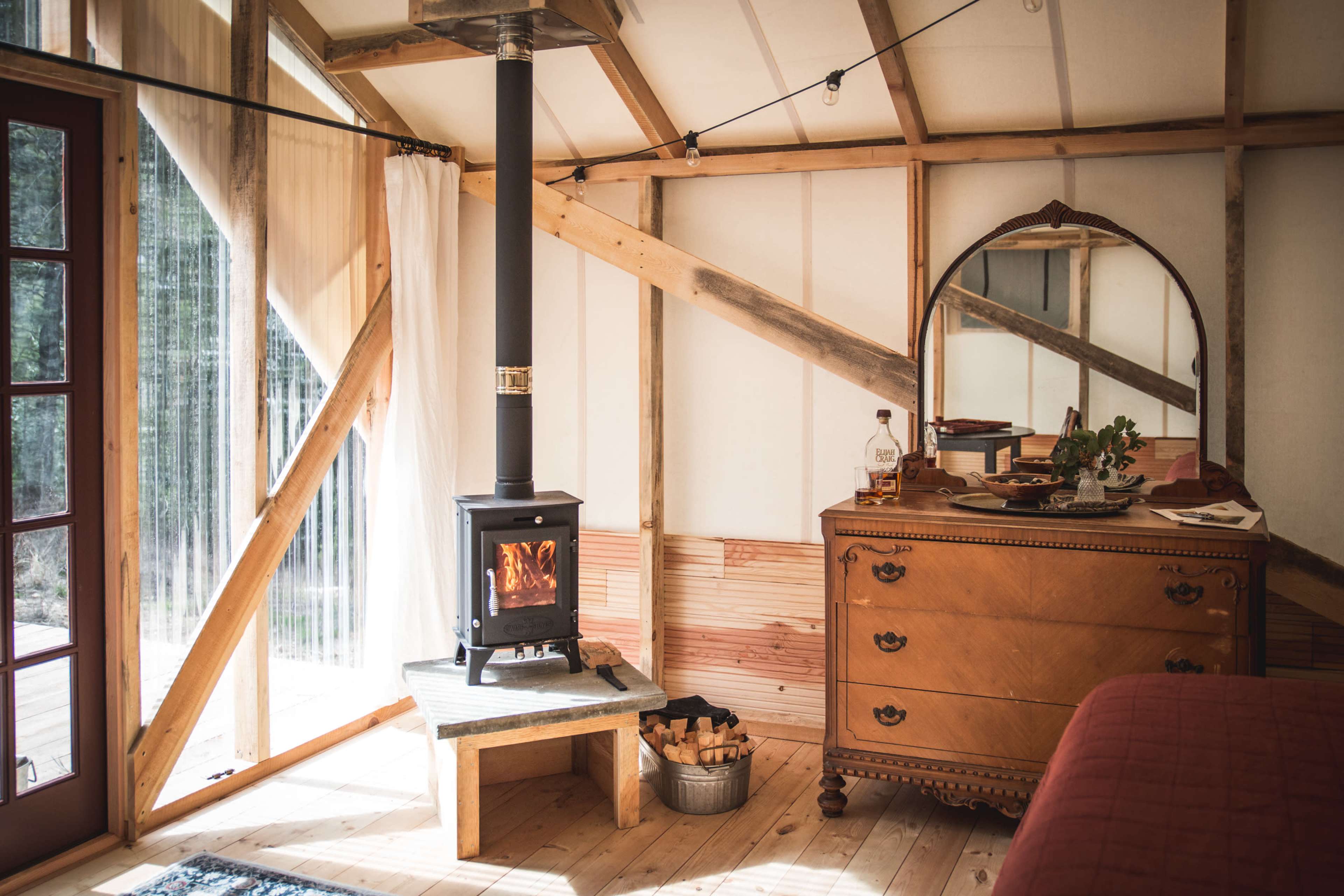 The interior of a cozy yurt features a wood stove, a vintage dresser, and large windows allowing natural light to enter.