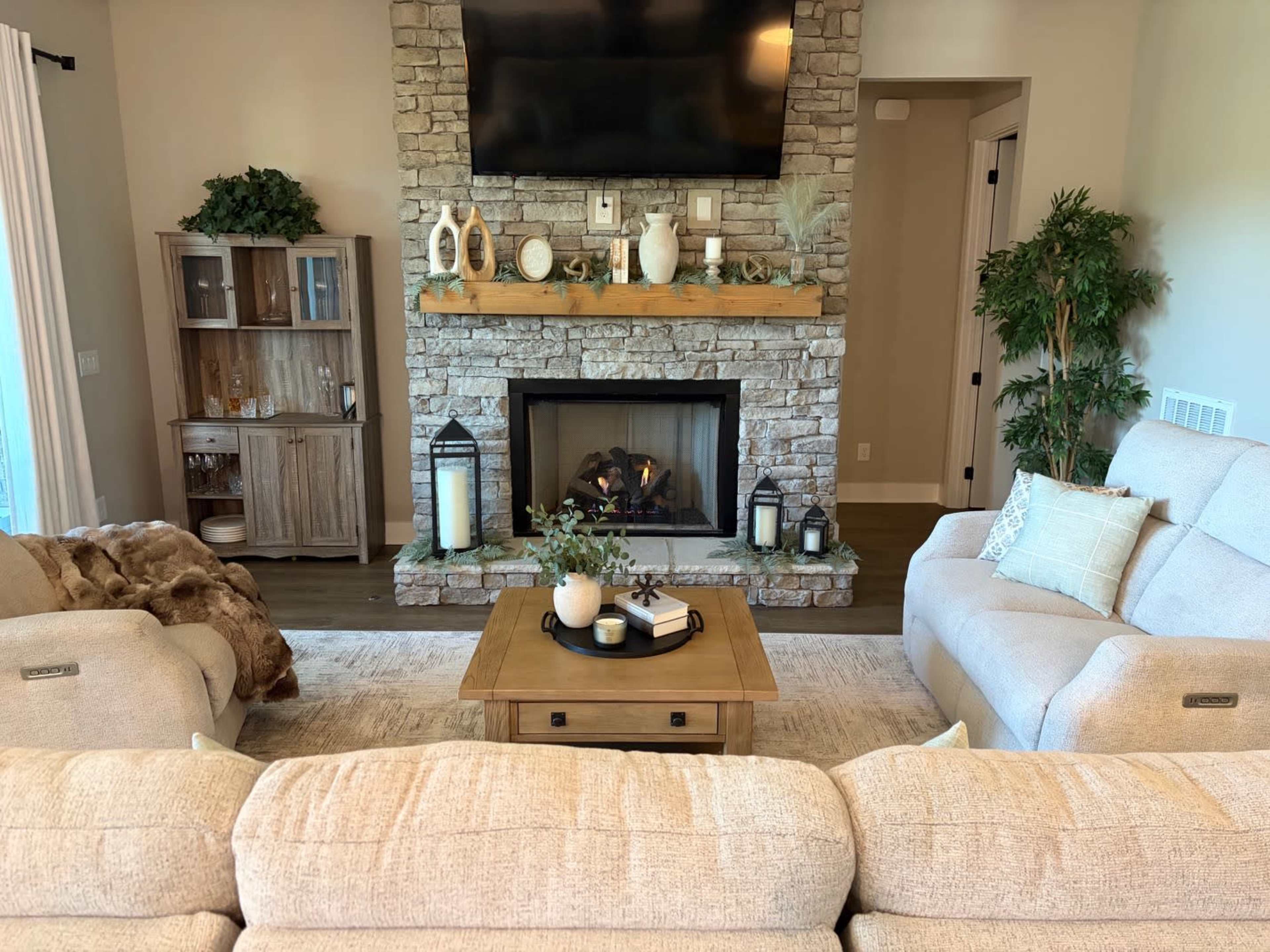 The image shows a cozy living room featuring a stone fireplace with a wood mantle, a large television mounted above it, and comfortable seating arranged around a central coffee table.
