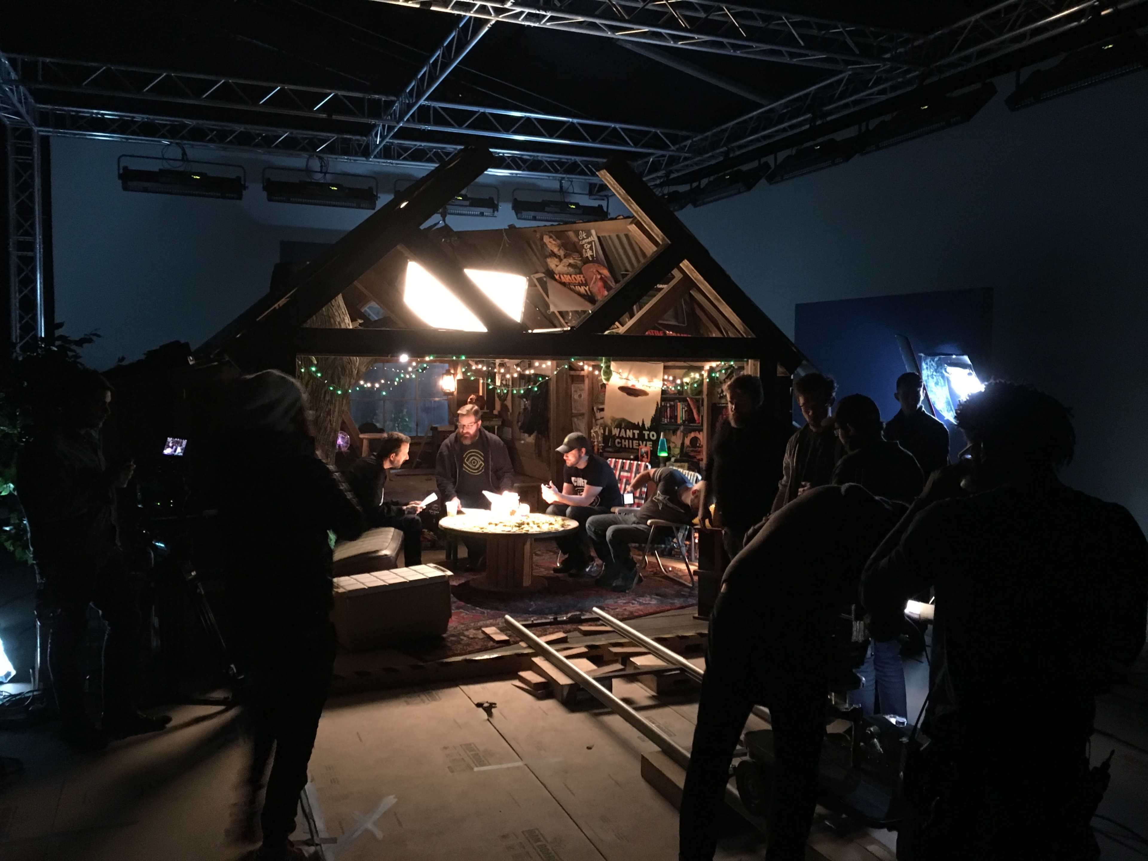 A group of people is gathered around a table inside a staged set designed to resemble a small cabin, with cameras and lights set up for filming.