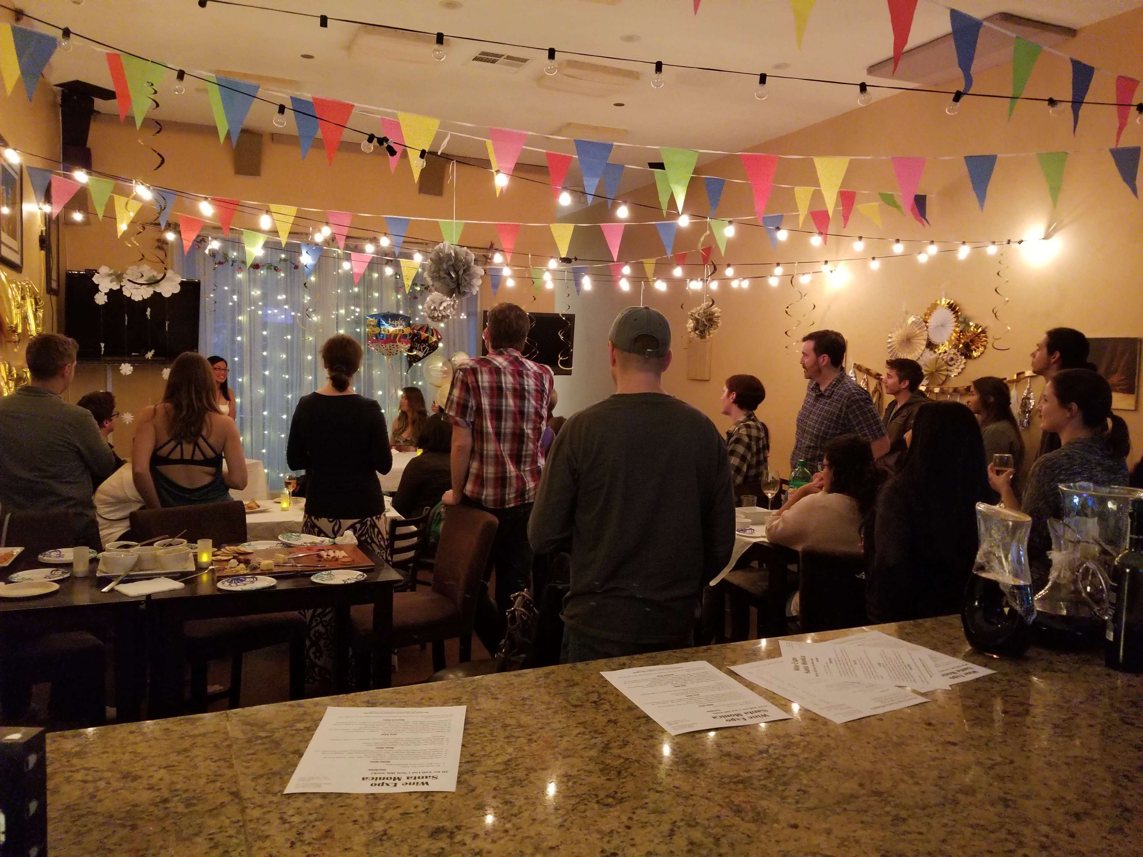 A group of people stands in a decorated room with colorful bunting and string lights, gathered around for an event.