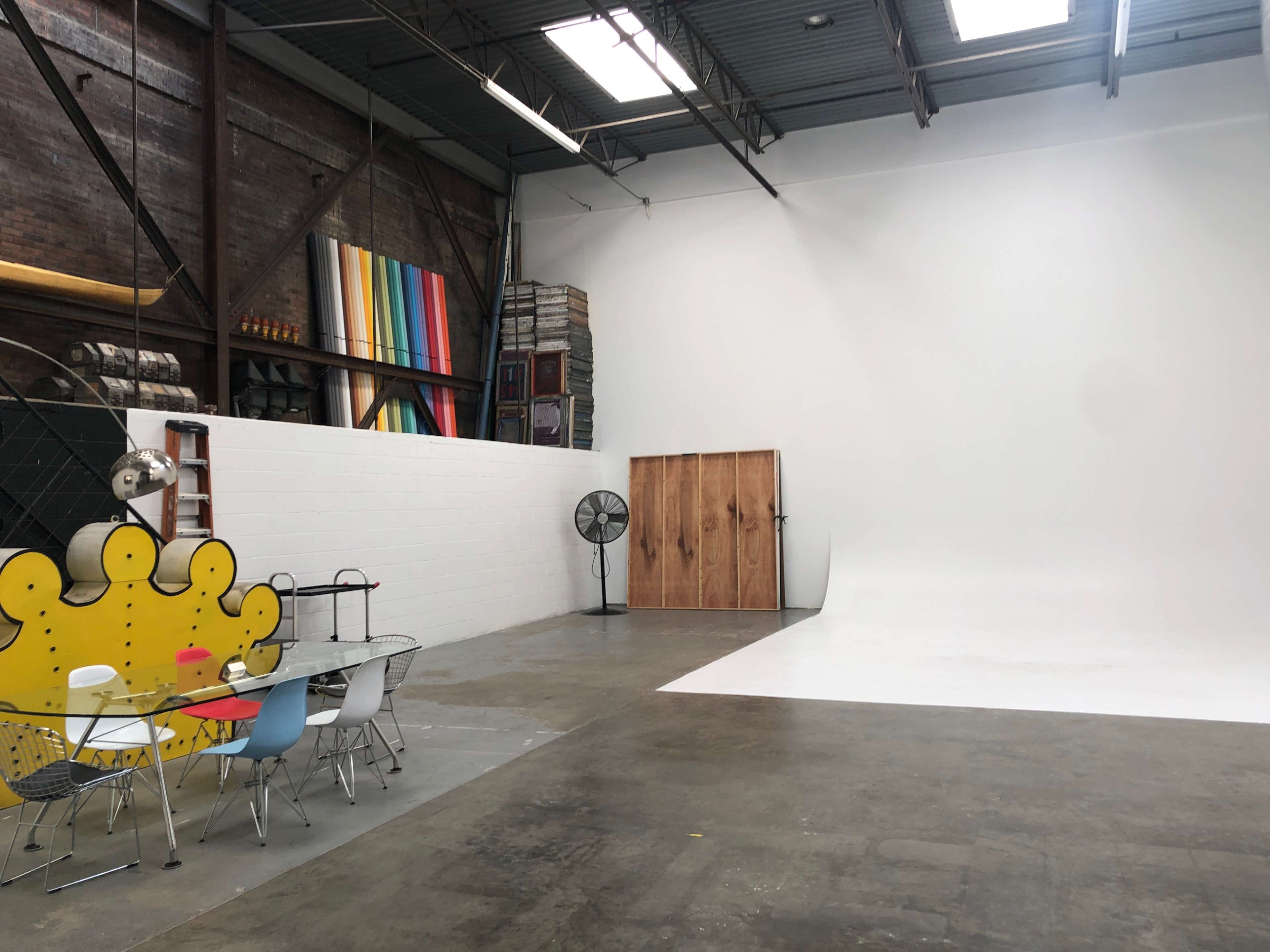 A spacious photography studio with a white backdrop, wooden door, colorful panels, and a table with chairs on a concrete floor.