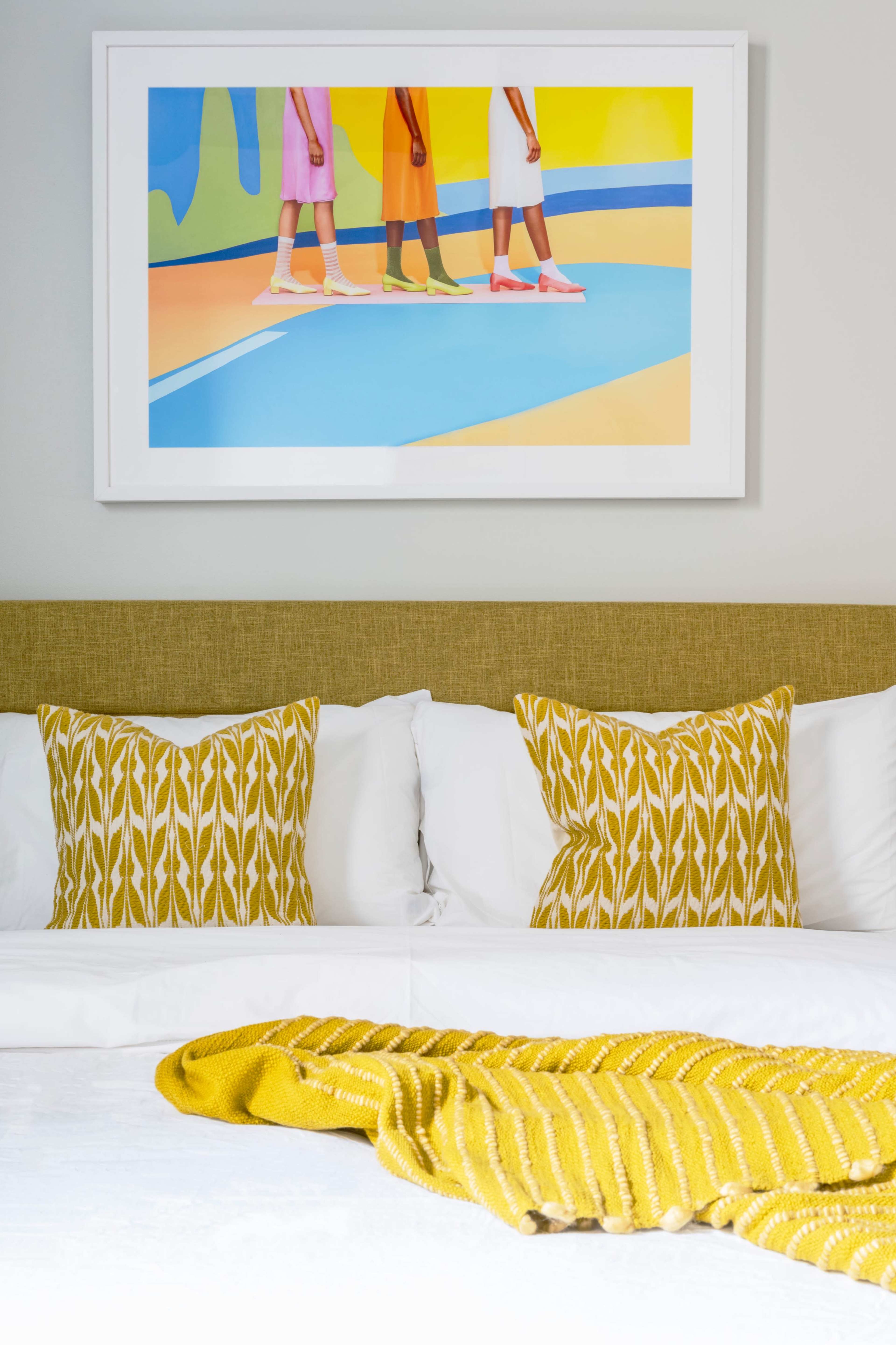 The image shows a styled bedroom featuring a neatly made bed with white linens, yellow patterned pillows, and a vibrant artwork of colorful figures on the wall above.