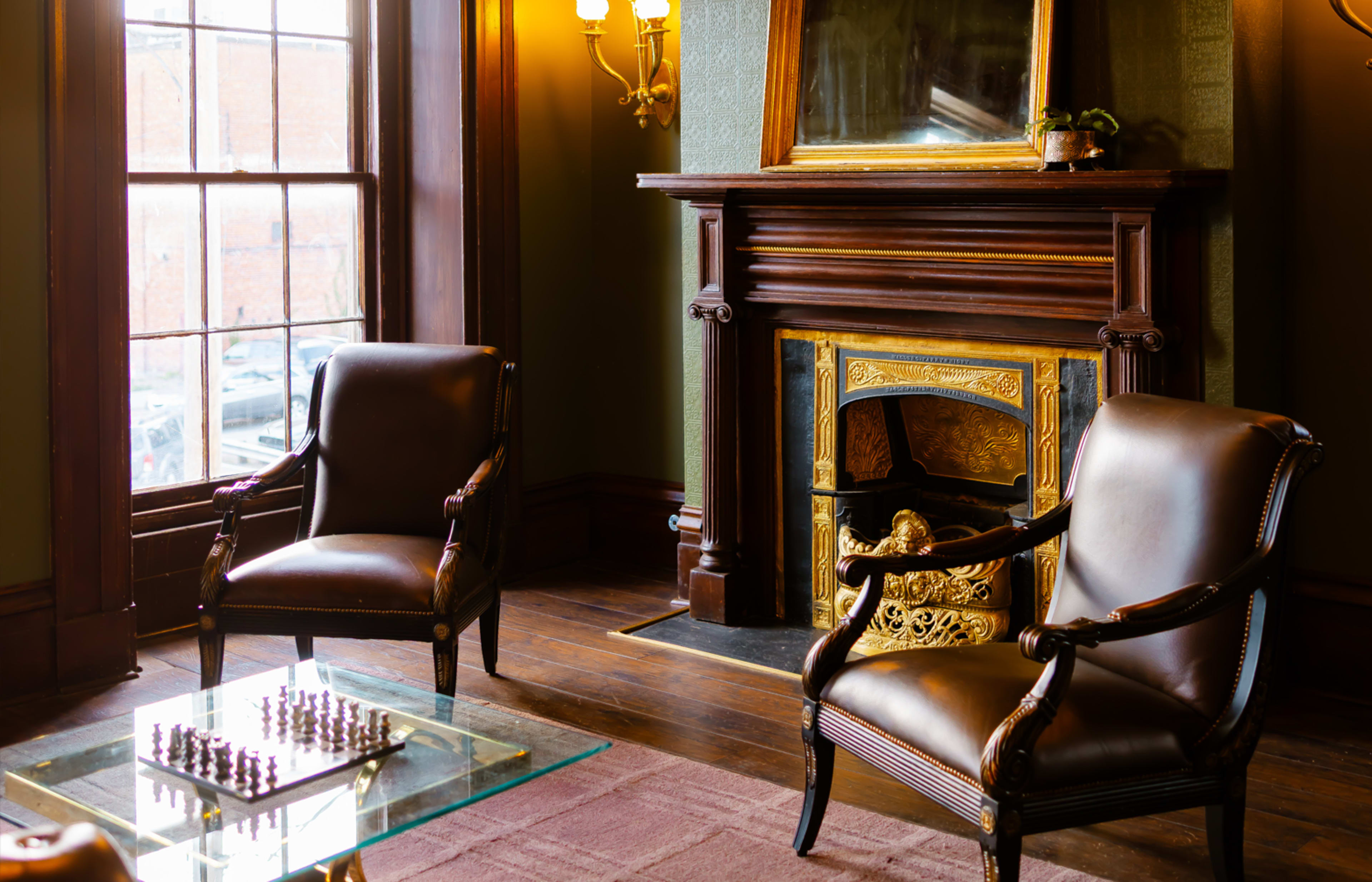 The image shows a cozy room with two leather armchairs, a glass coffee table with a chessboard, and a decorative fireplace with a mirror above it.