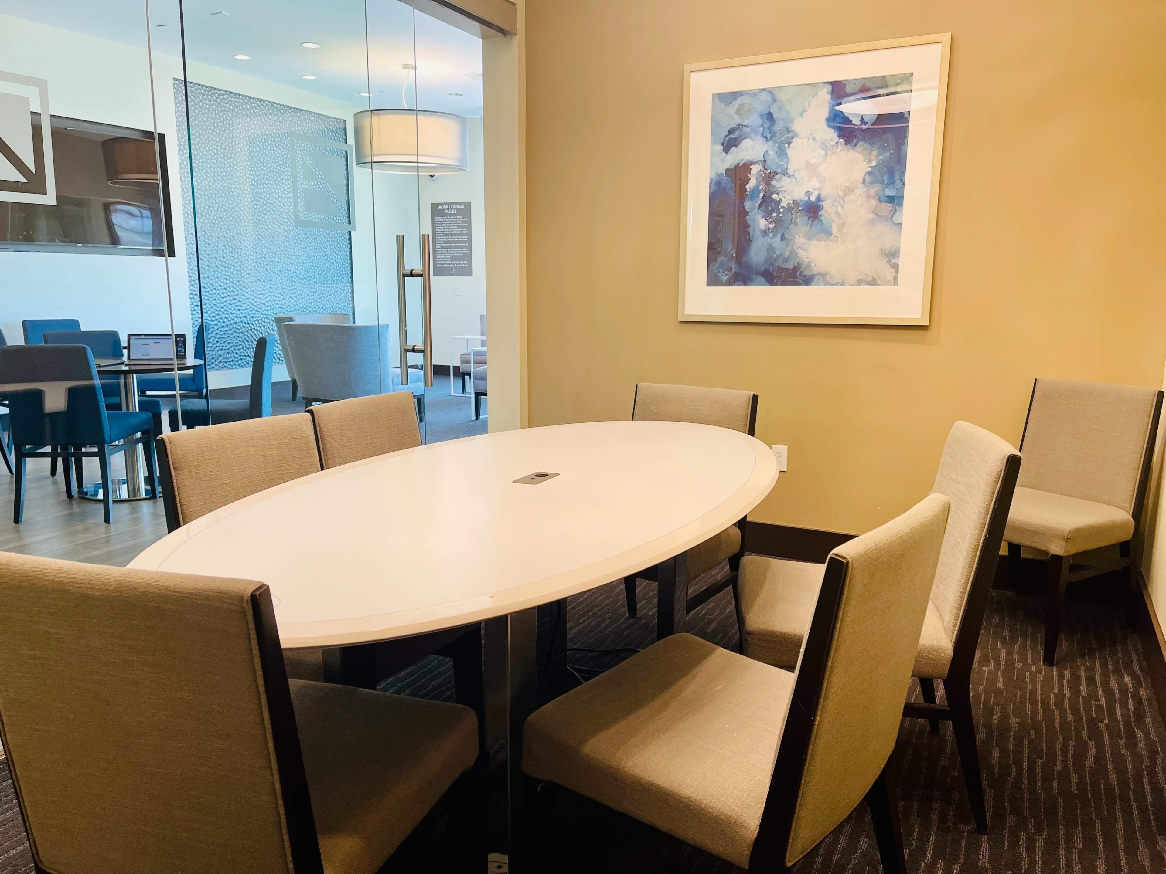 A round table surrounded by six chairs is situated in a meeting room with a framed abstract painting on the wall.