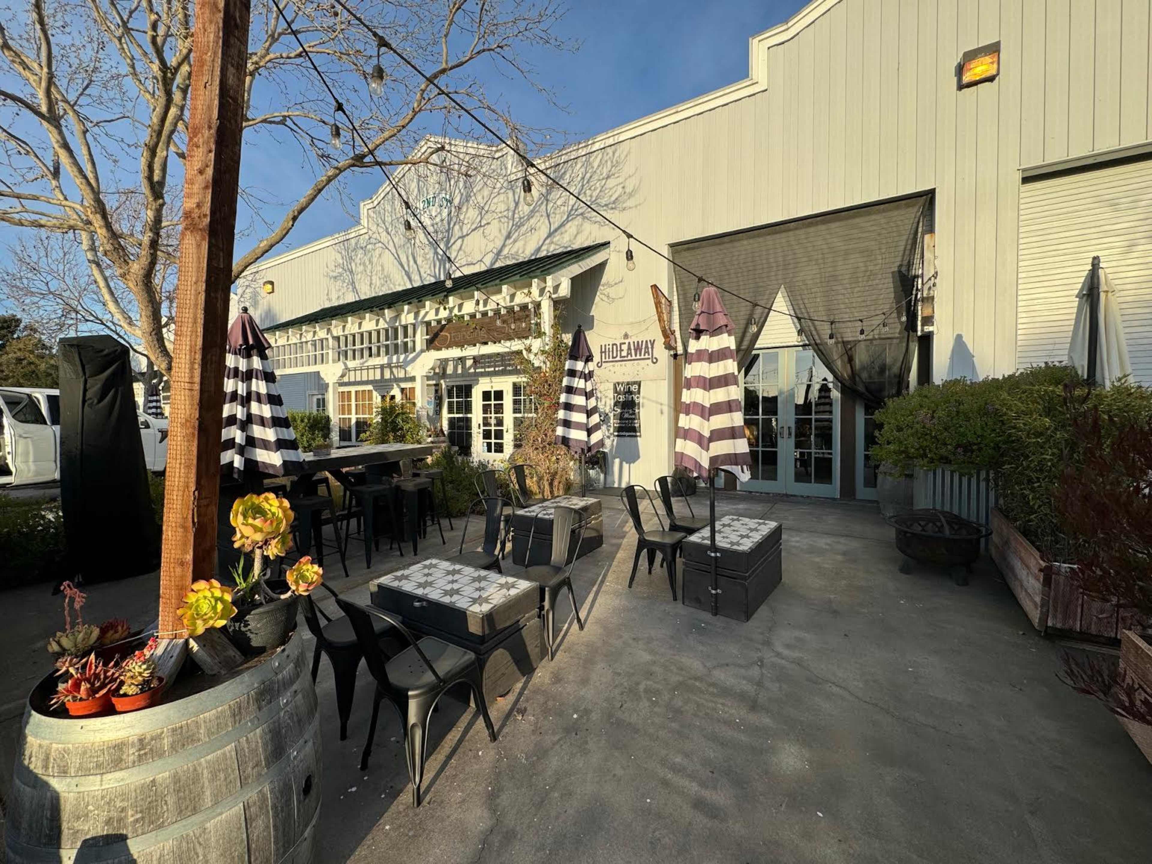 An outdoor seating area of a restaurant with striped umbrellas, black chairs, and potted plants in front of a building.