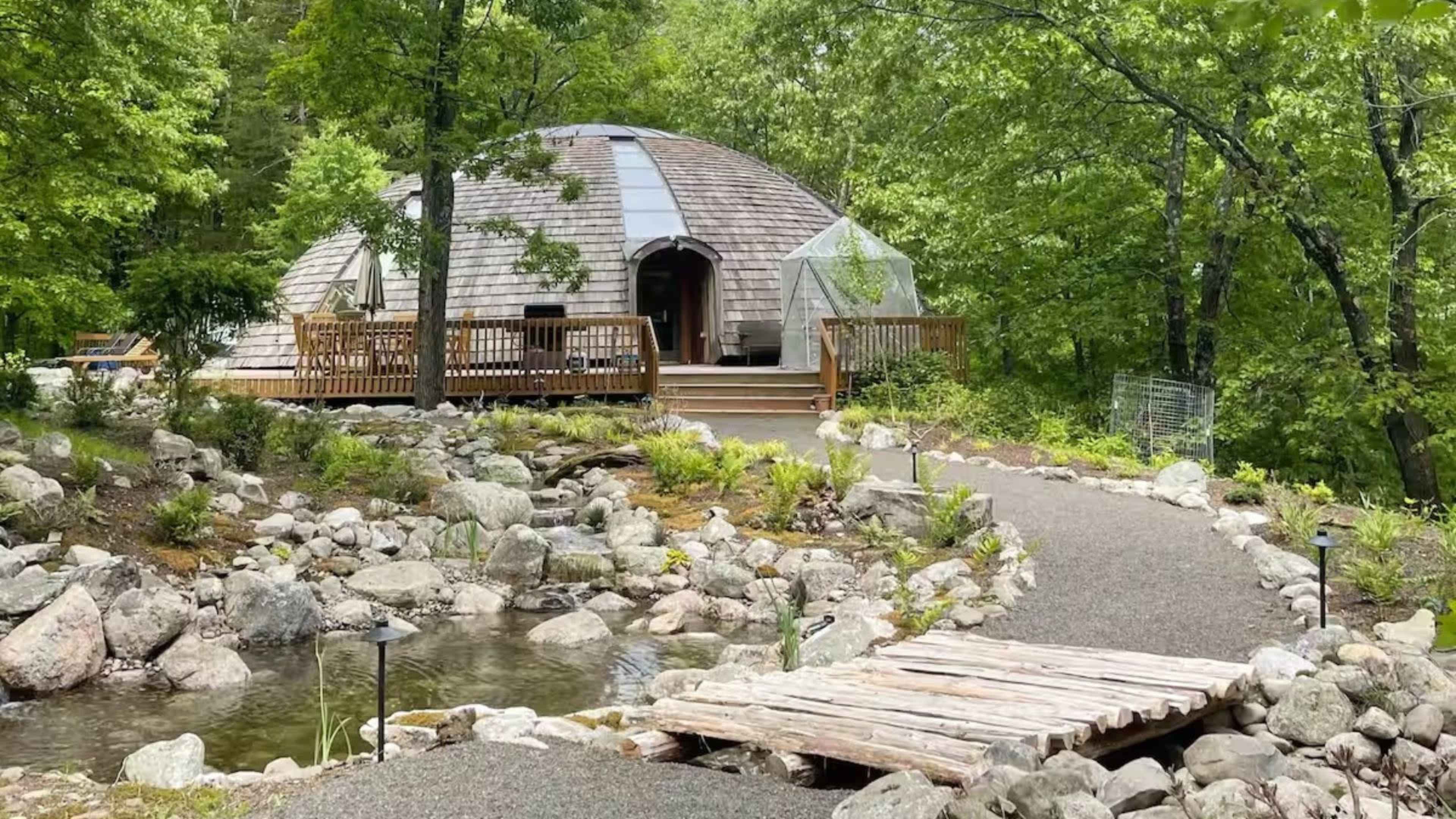 A circular wooden structure with a shingled roof is surrounded by lush greenery and a pond, connected to a path by a small wooden bridge.