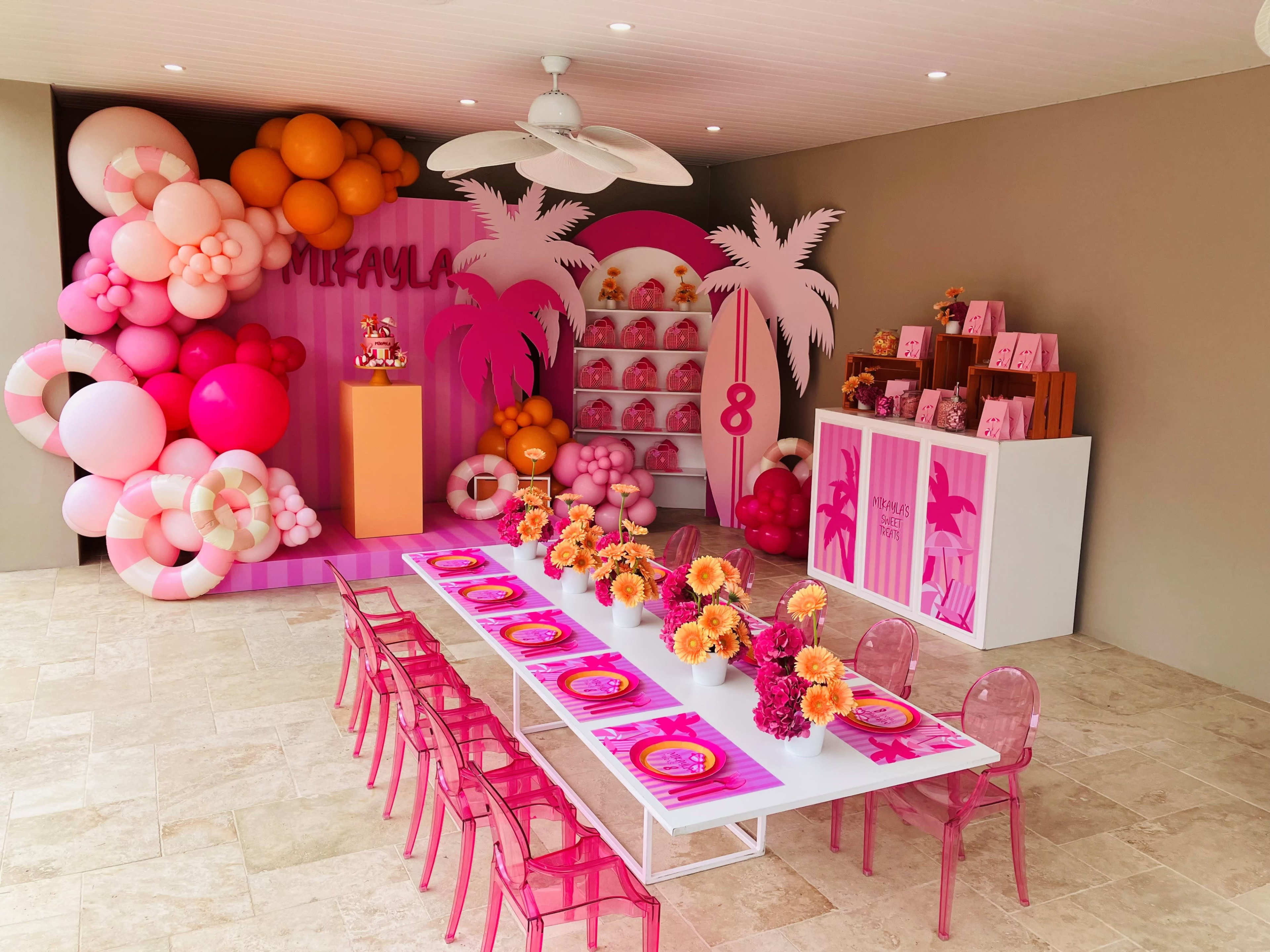 A colorful party setup features a decorated table with pink chairs, vibrant floral arrangements, and a balloon arch backdrop, creating a festive atmosphere.