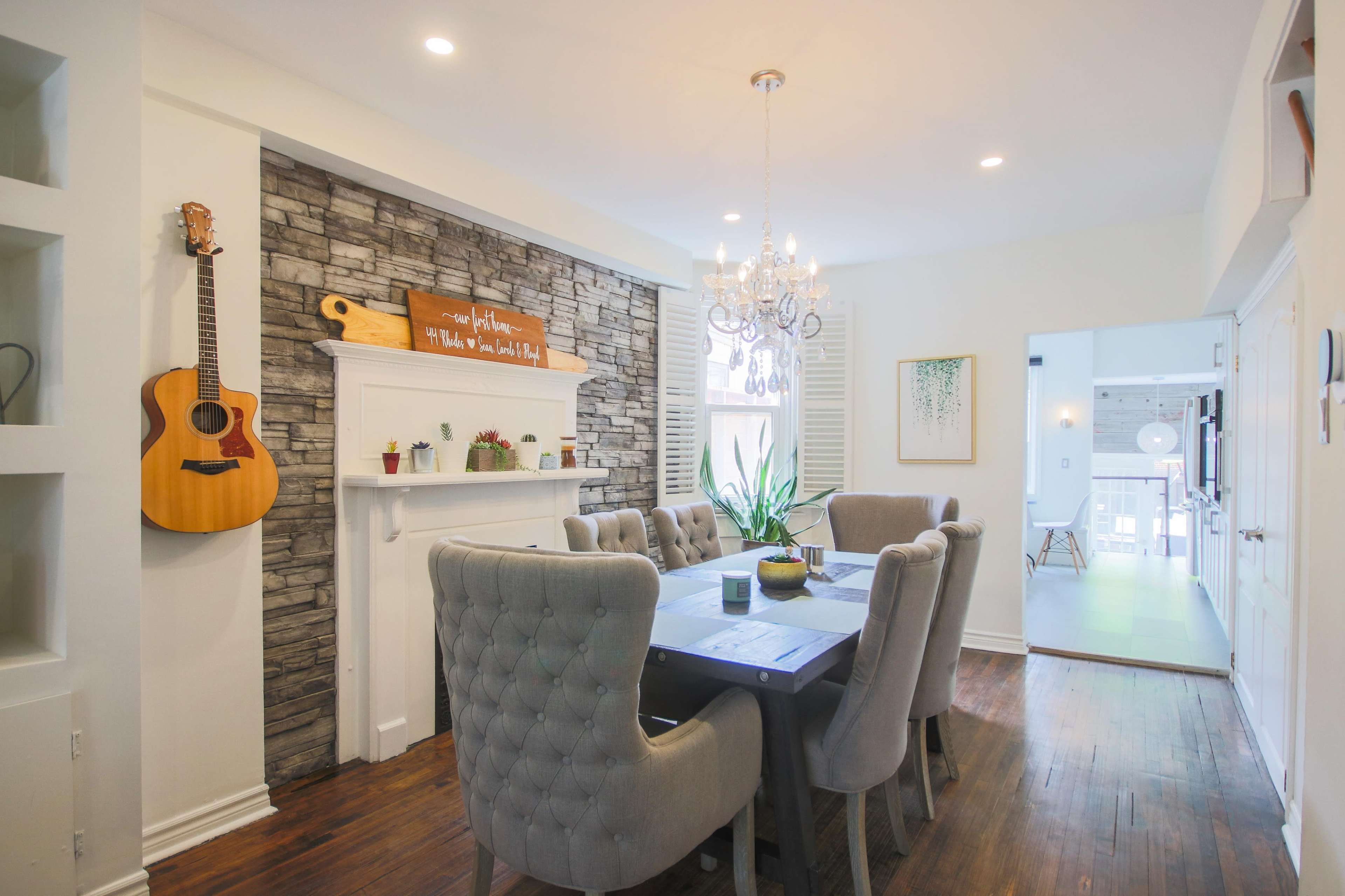 A dining area features a stone accent wall, a large gray table surrounded by upholstered chairs, and a chandelier above, with a guitar hanging on the wall.
