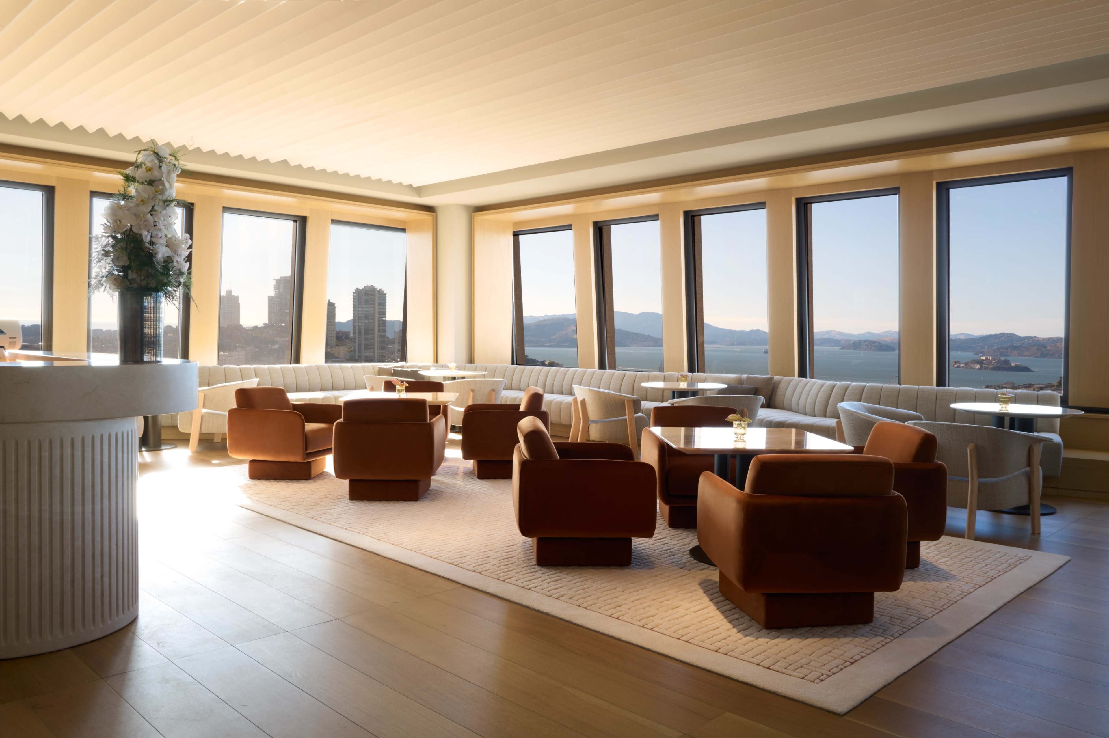 The image shows a modern lounge area with large windows, featuring comfortable brown chairs and tables against a backdrop of a city skyline and mountains.
