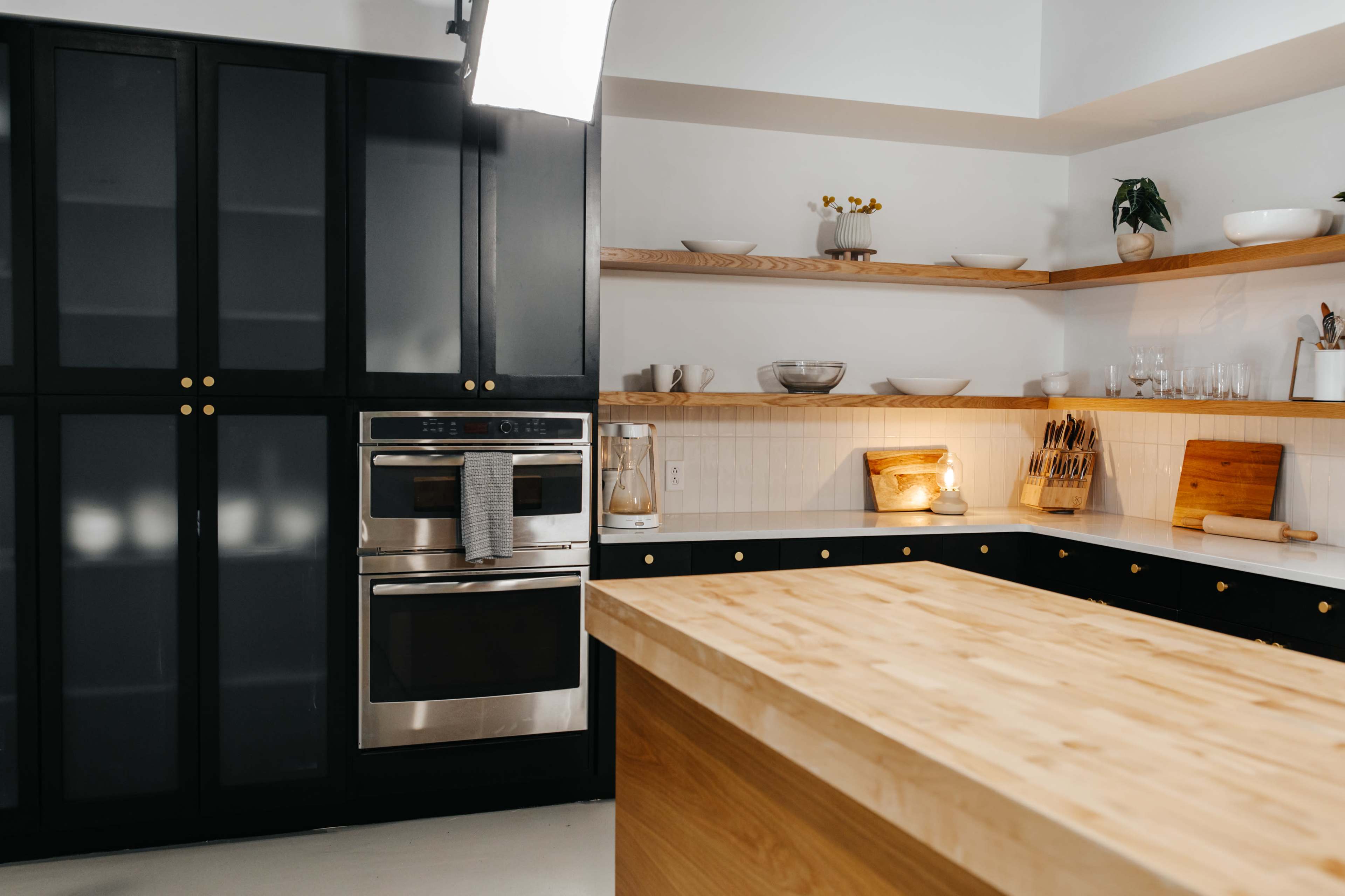 A modern kitchen features black cabinetry, stainless steel appliances, and a large wooden island with shelves displaying dishware.