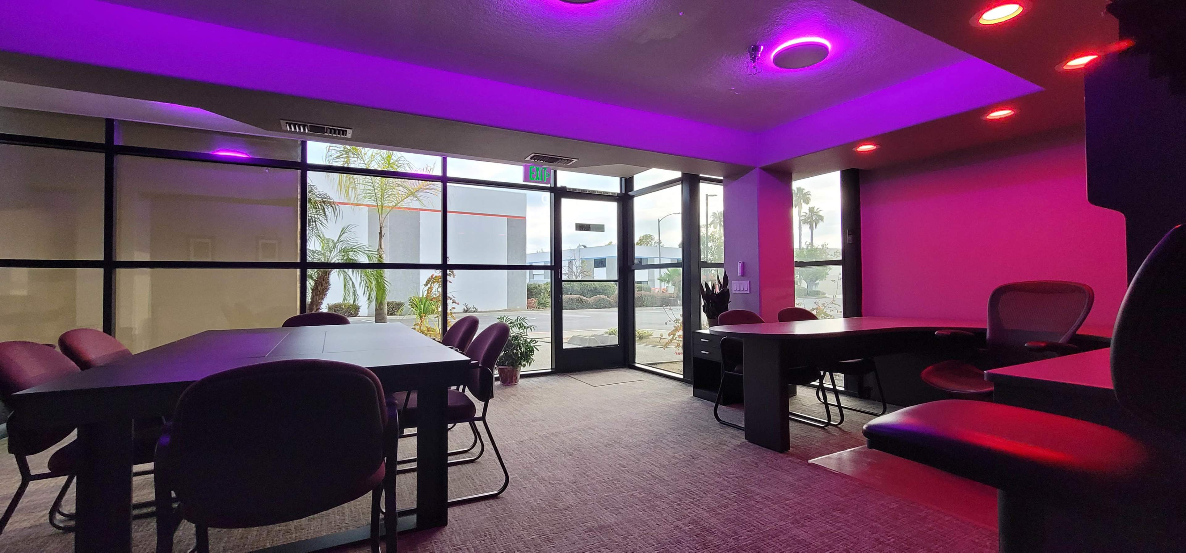 A modern conference room features dark tables and chairs, illuminated by purple and red lights, with large glass windows showing a view of palm trees outside.