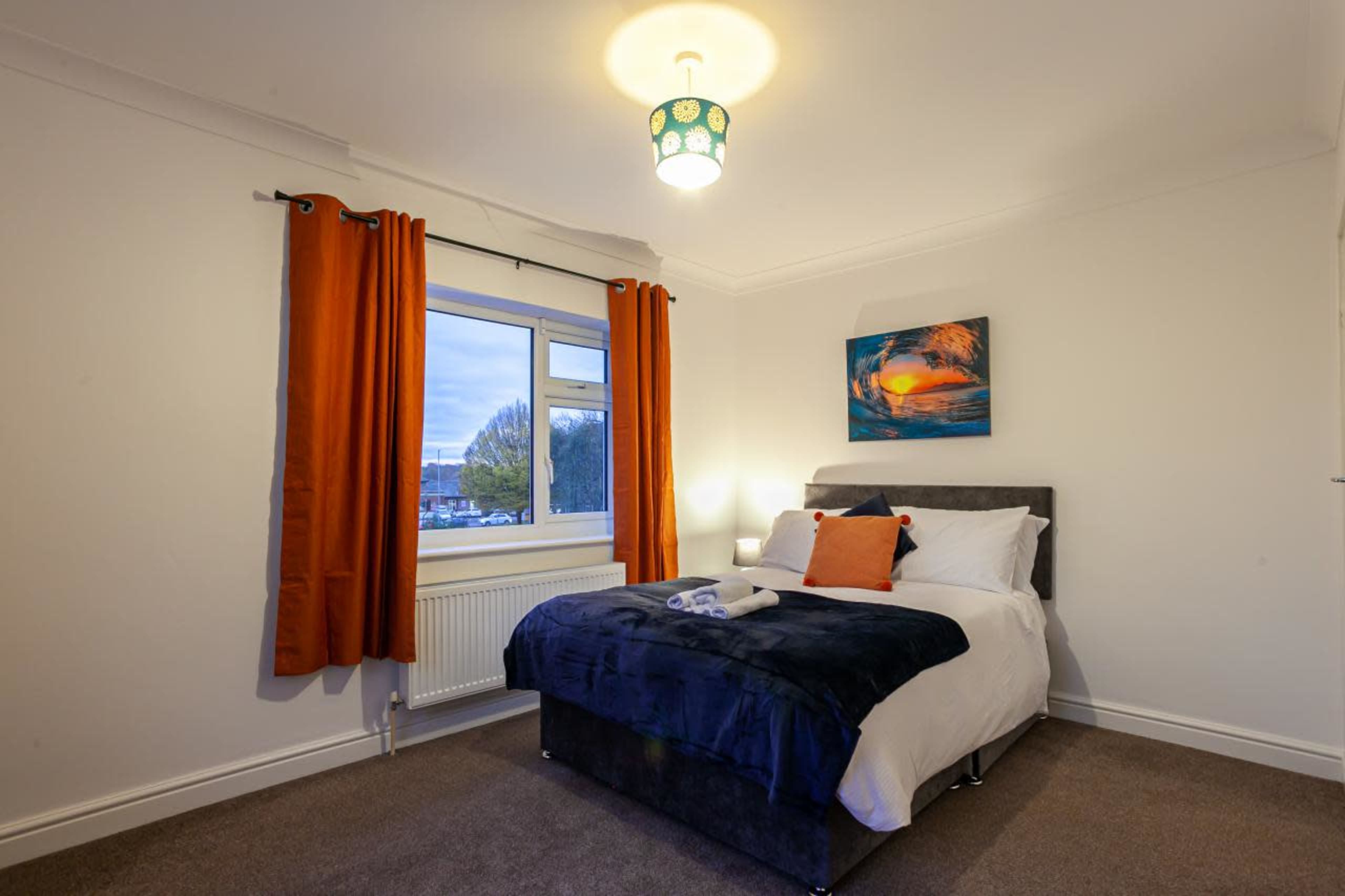 A neat bedroom with a double bed, orange curtains, and a colorful wall art piece, along with a window providing natural light.