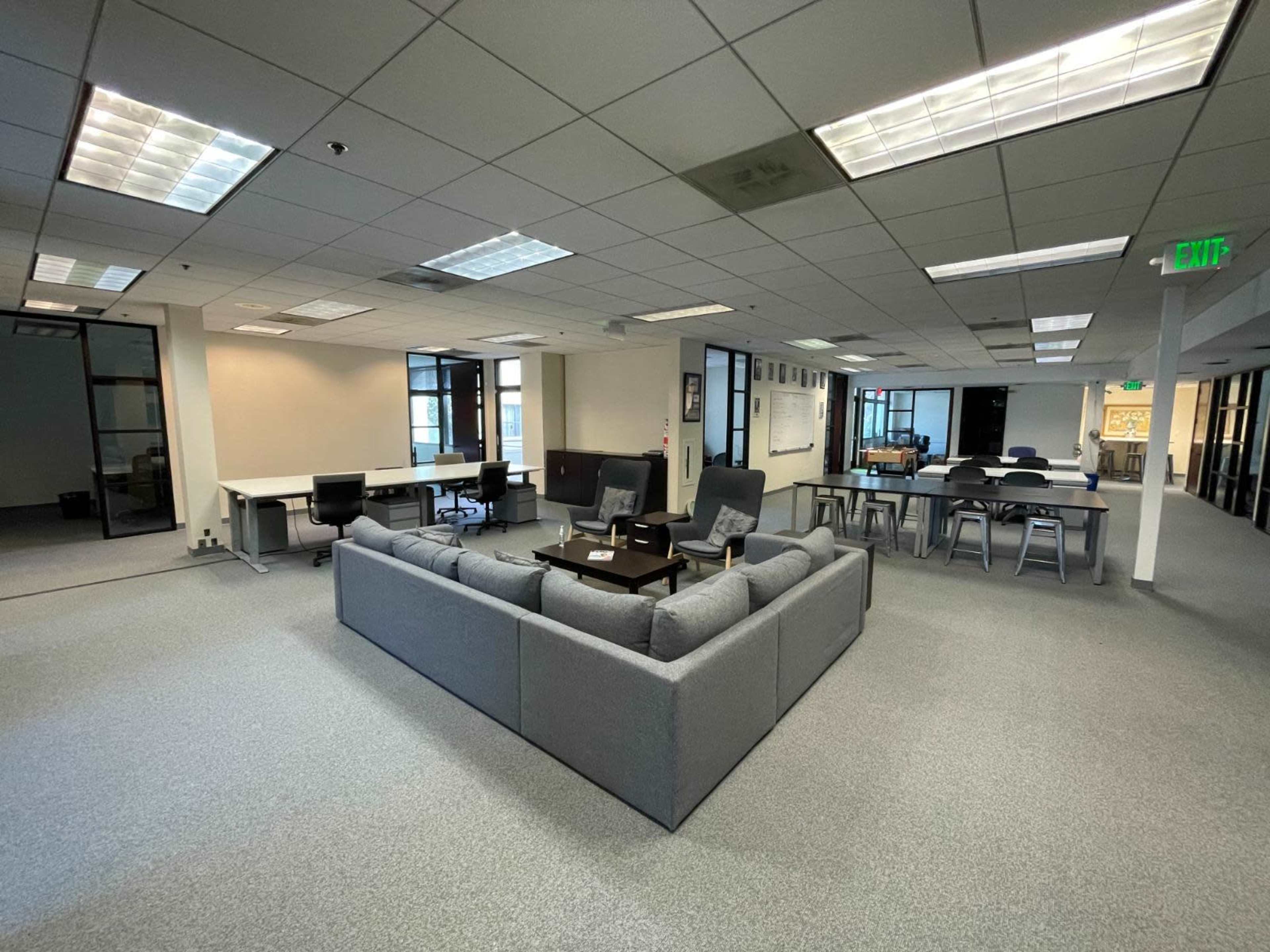 An office space with a large gray sofa arrangement in the center, surrounded by desks and chairs, and equipped with large windows.