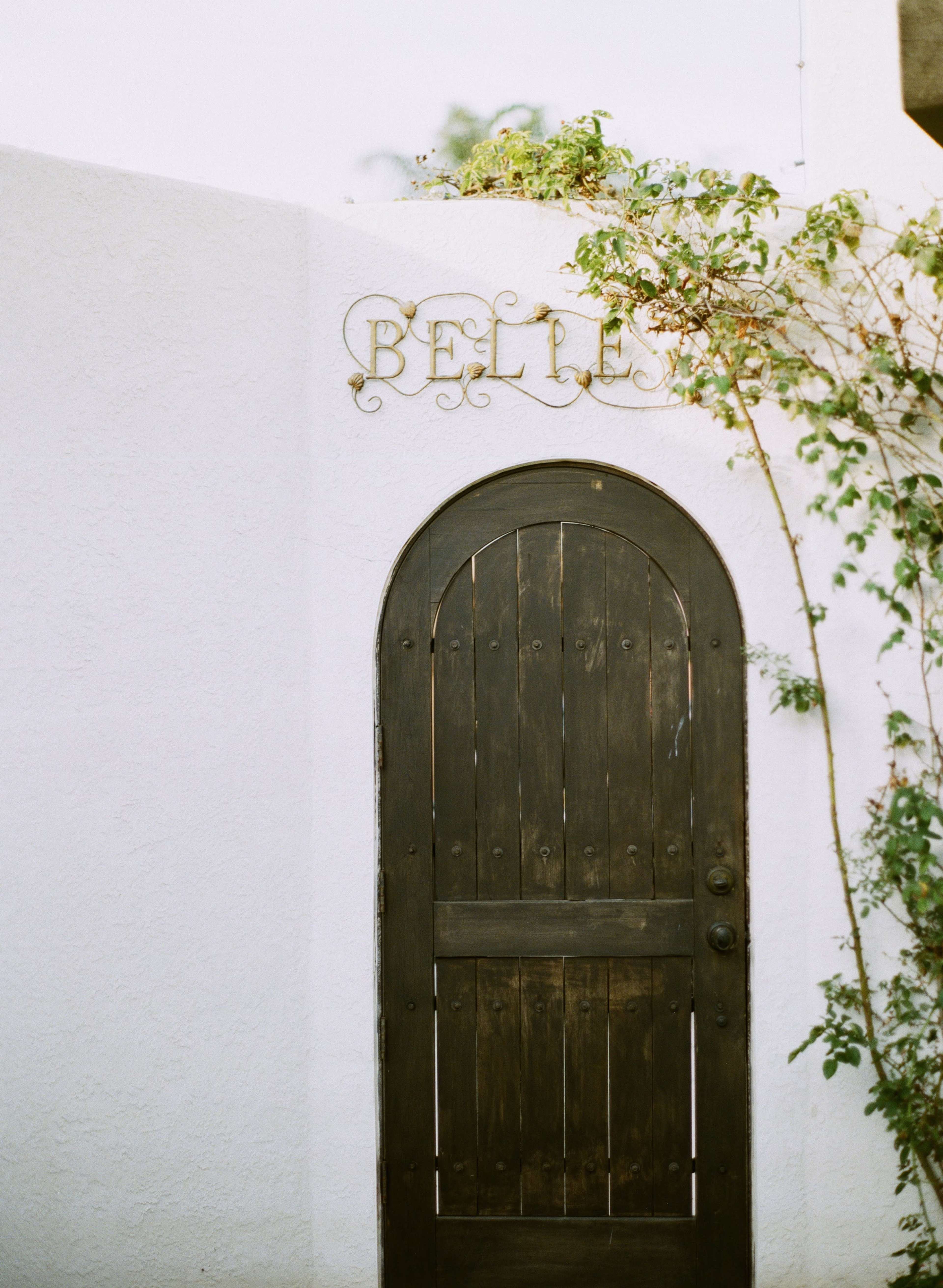 The image features an arched wooden door with a weathered finish, set into a white wall, adorned with the word "BELLE" and flanked by climbing greenery.