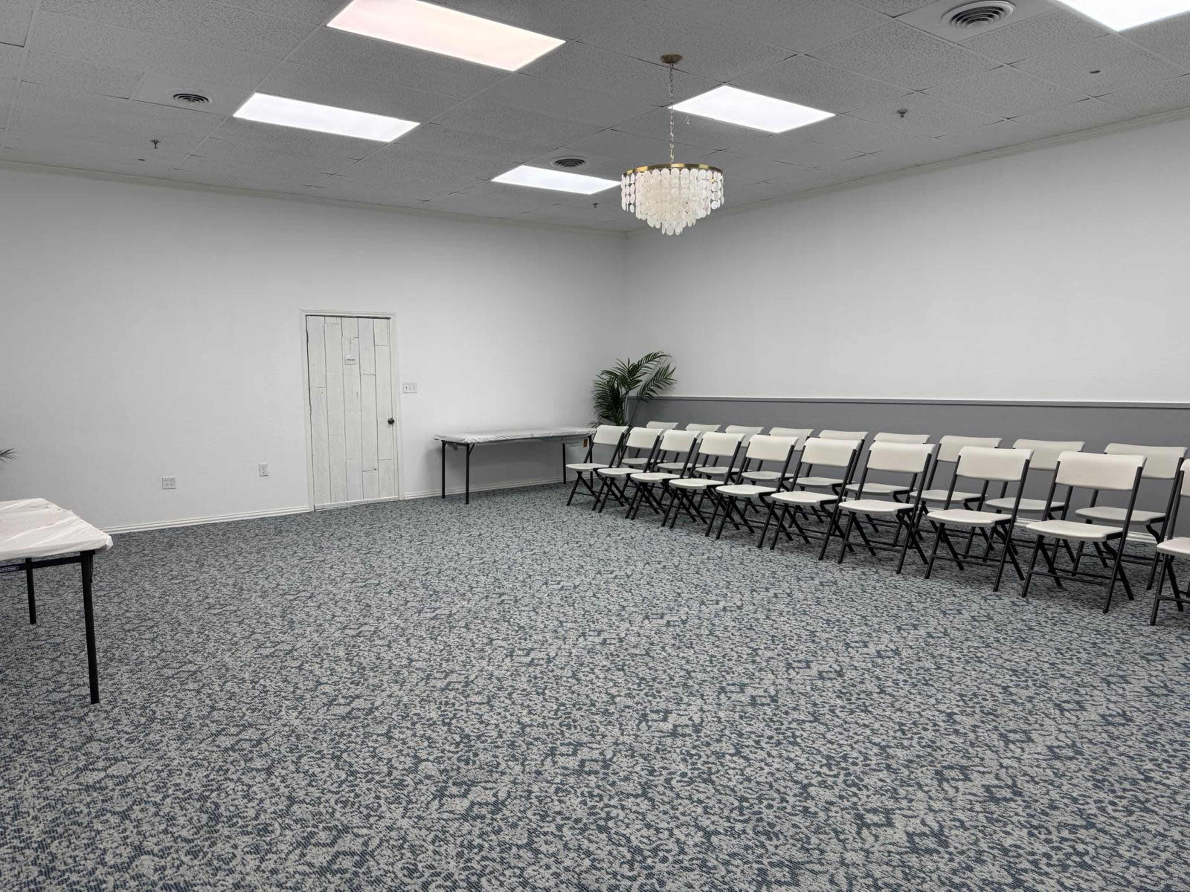 The image shows a sparsely furnished room with a patterned carpet, a chandelier, a door, and several rows of white folding chairs set up for an event.