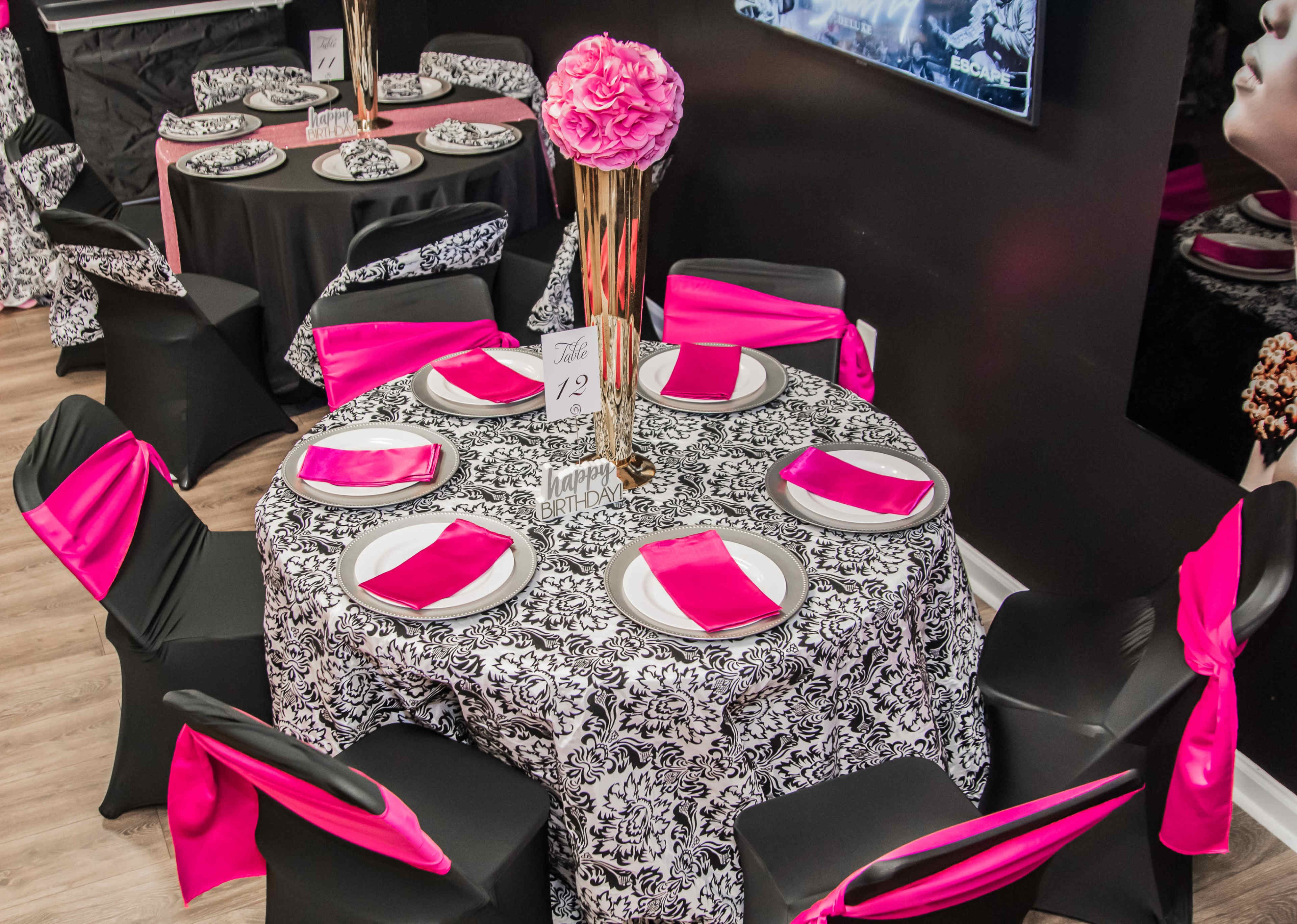 A dining area is set up with black and white patterned tablecloths, pink napkins, and tall centerpieces on each table.
