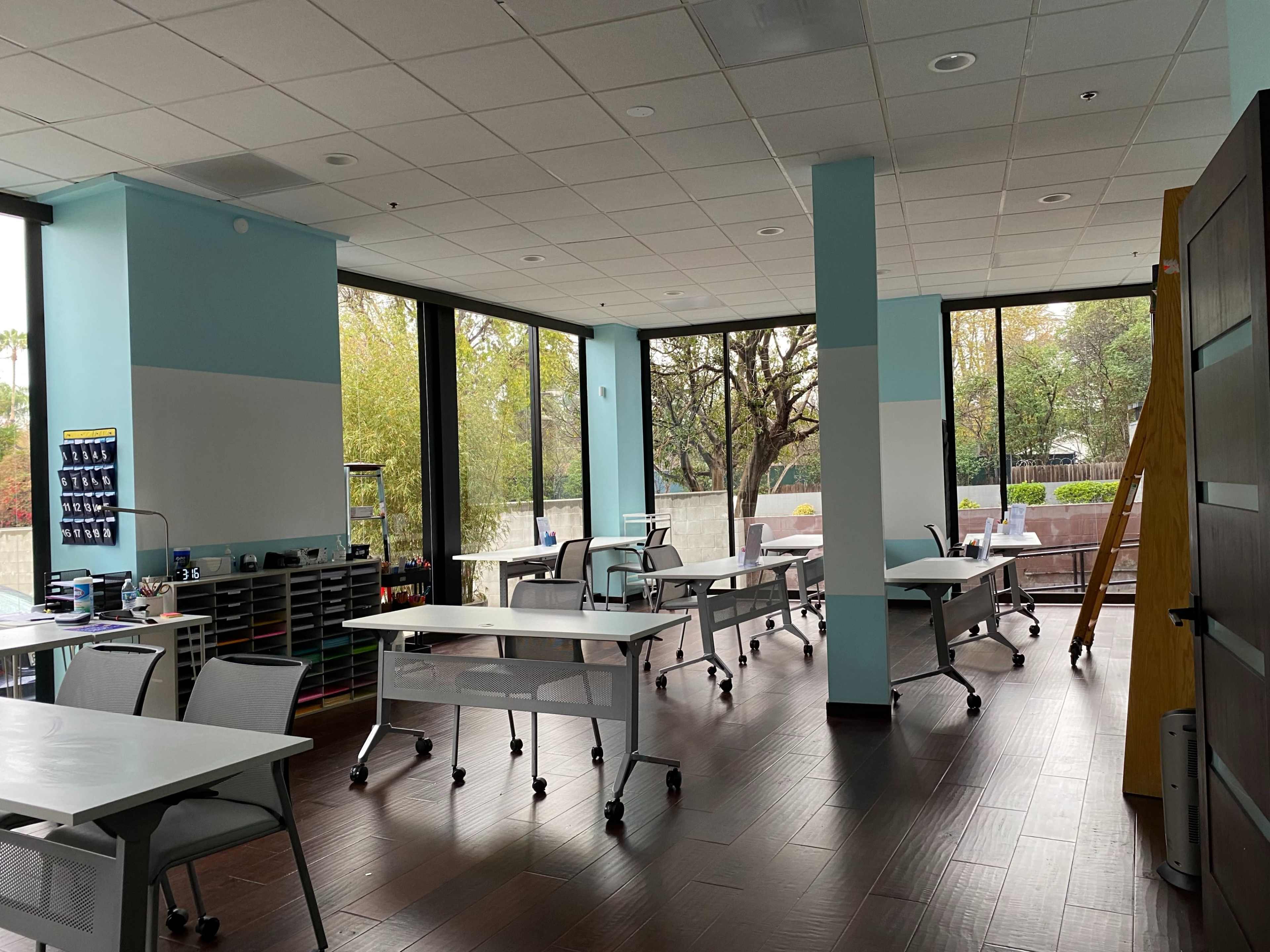 A spacious classroom features several desks arranged throughout, large windows allowing natural light, and storage areas along the walls.
