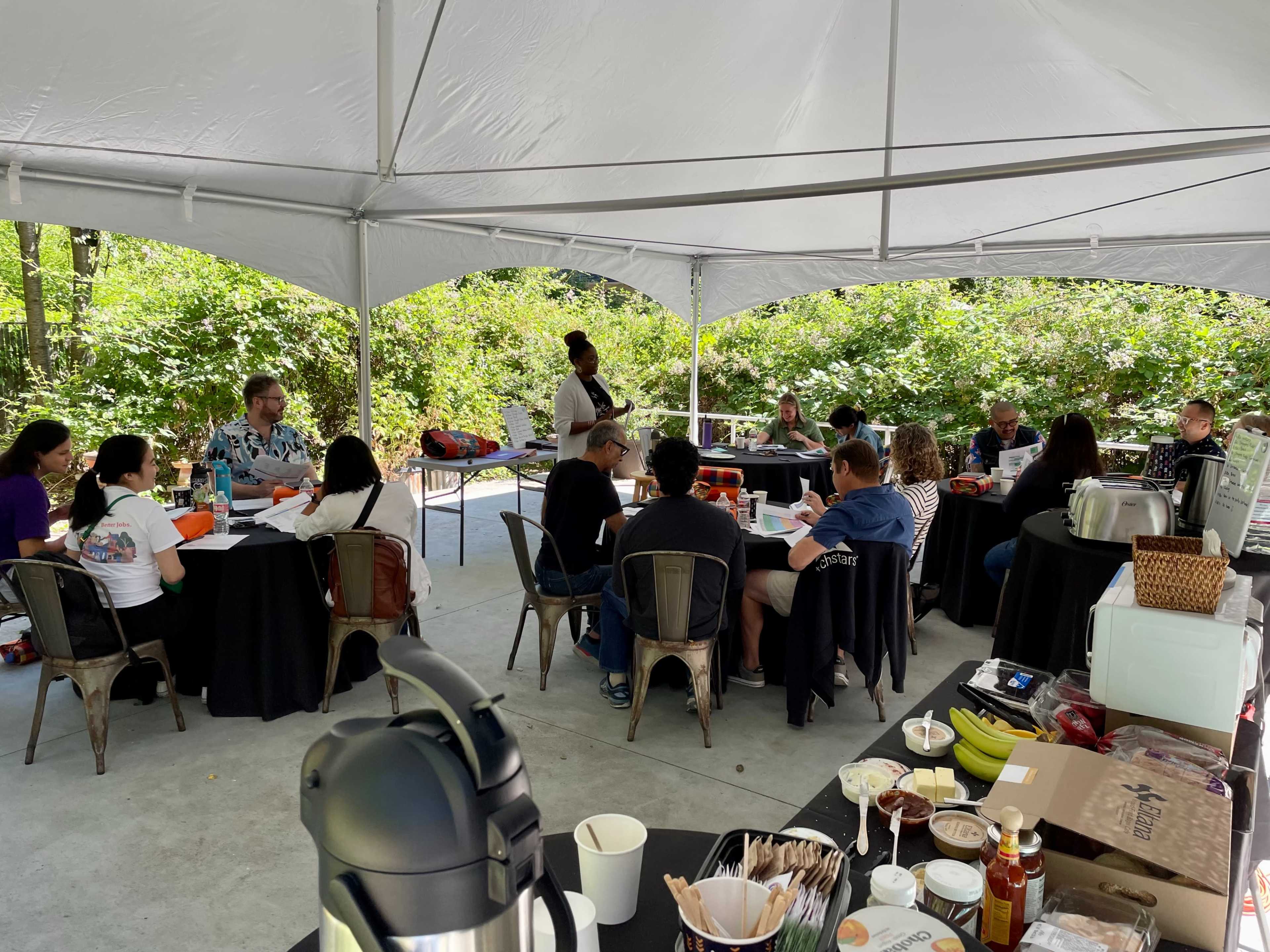 A group of people are seated around tables under a large tent, engaged in a discussion while food and beverages are set out on nearby tables.
