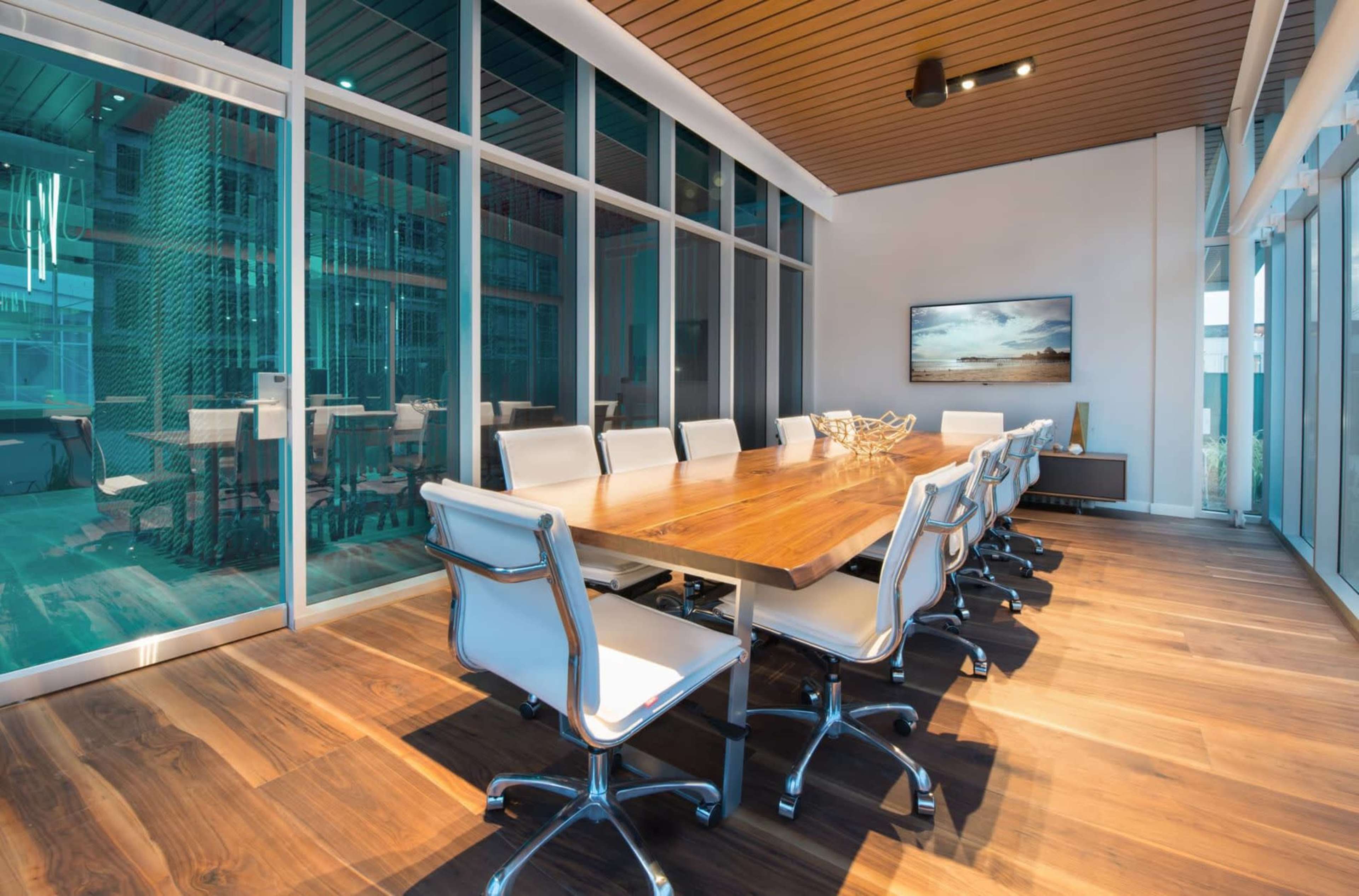 The image shows a modern conference room with a large wooden table surrounded by white chairs and glass walls, featuring a view of another meeting area.