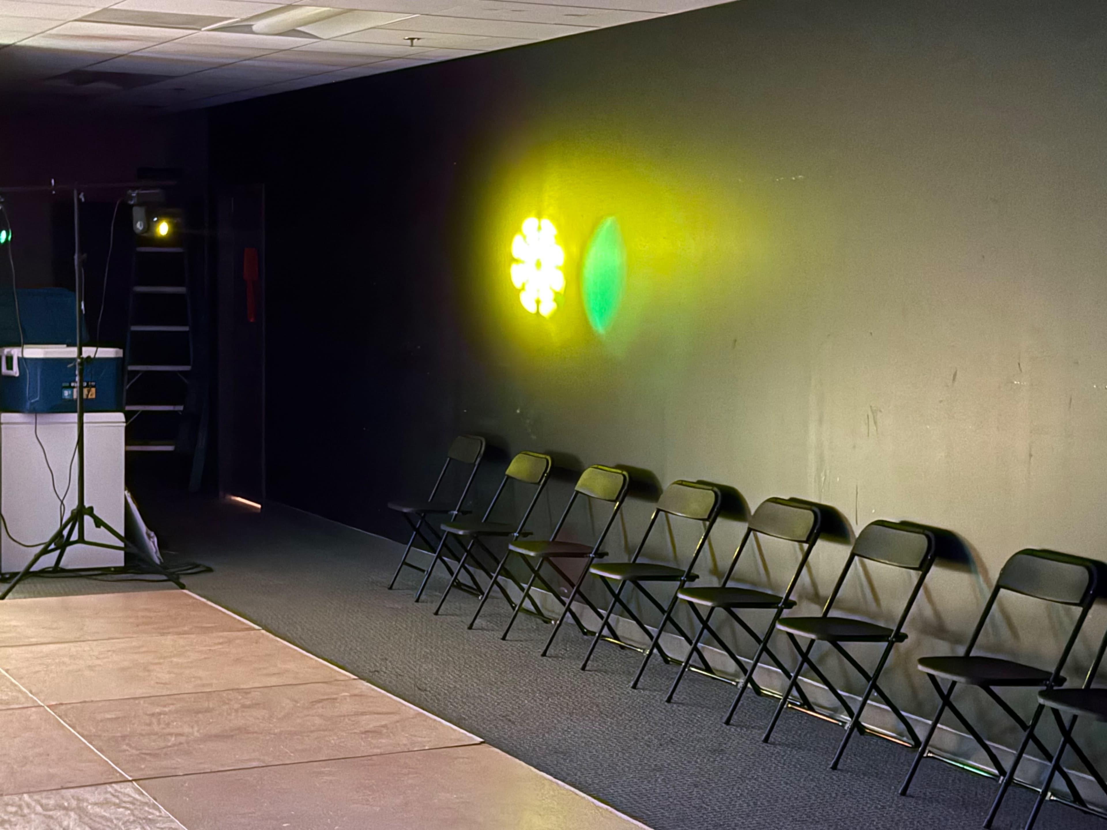 The image shows a row of black folding chairs against a dark wall, illuminated by colorful light spots.