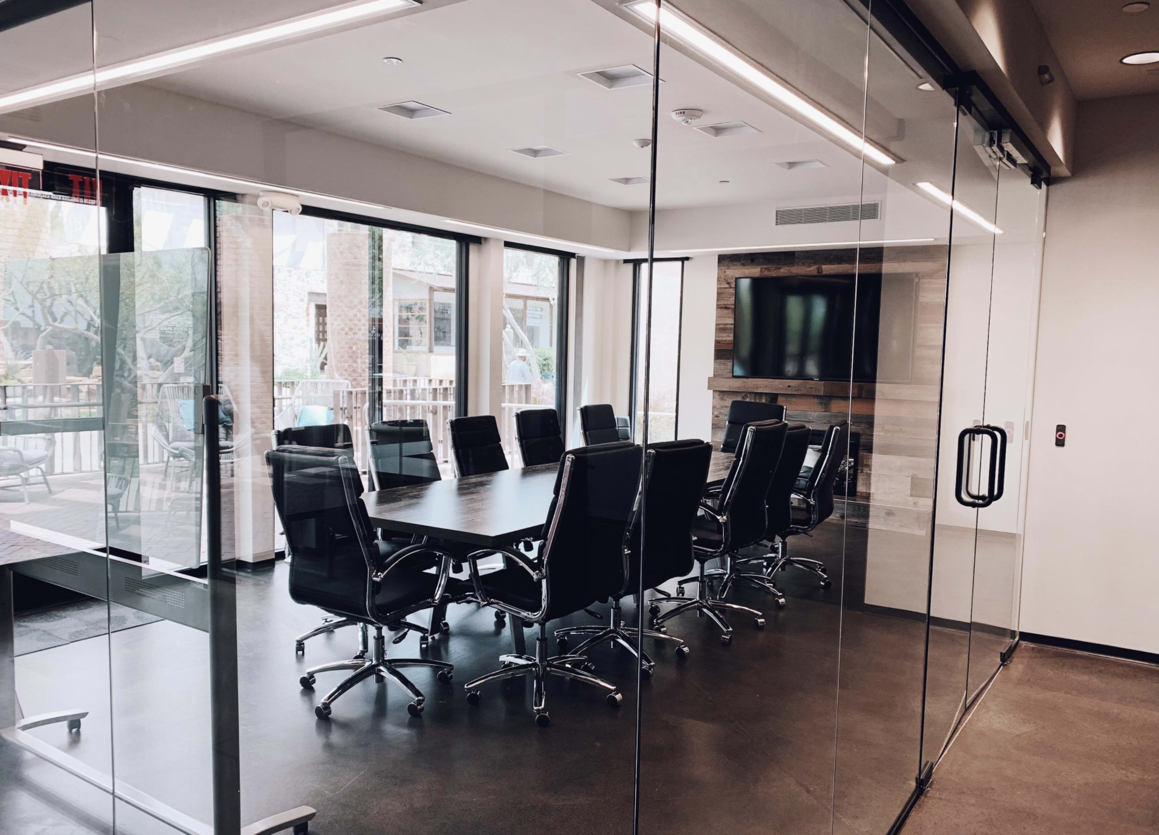 A large conference room features a long table surrounded by black leather chairs, with walls of glass providing a view of the outside.