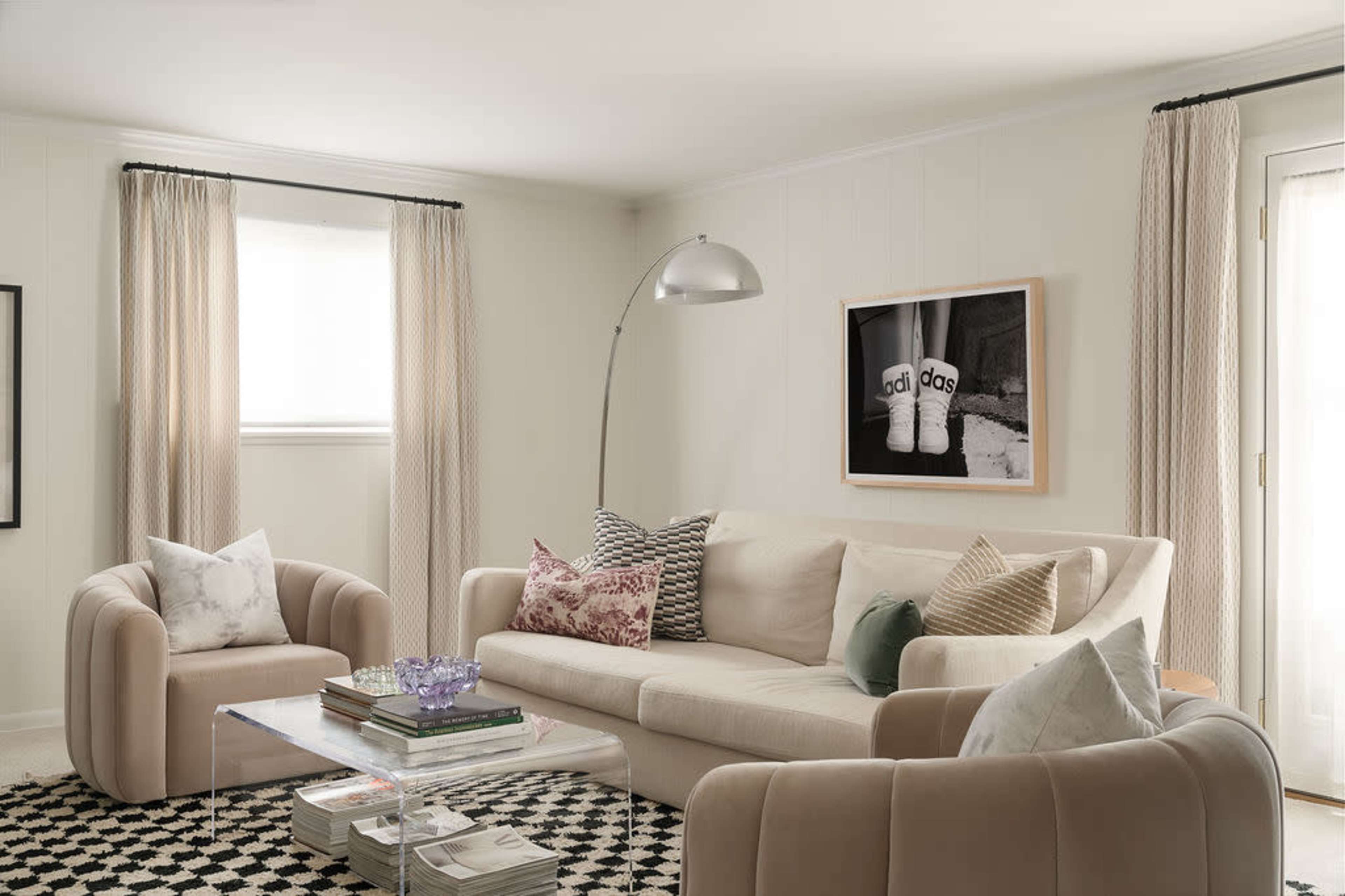 A modern living room with a light-colored sofa, patterned throw pillows, an arc floor lamp, and a coffee table topped with books and a decorative item, all set against a backdrop of neutral walls and large windows.