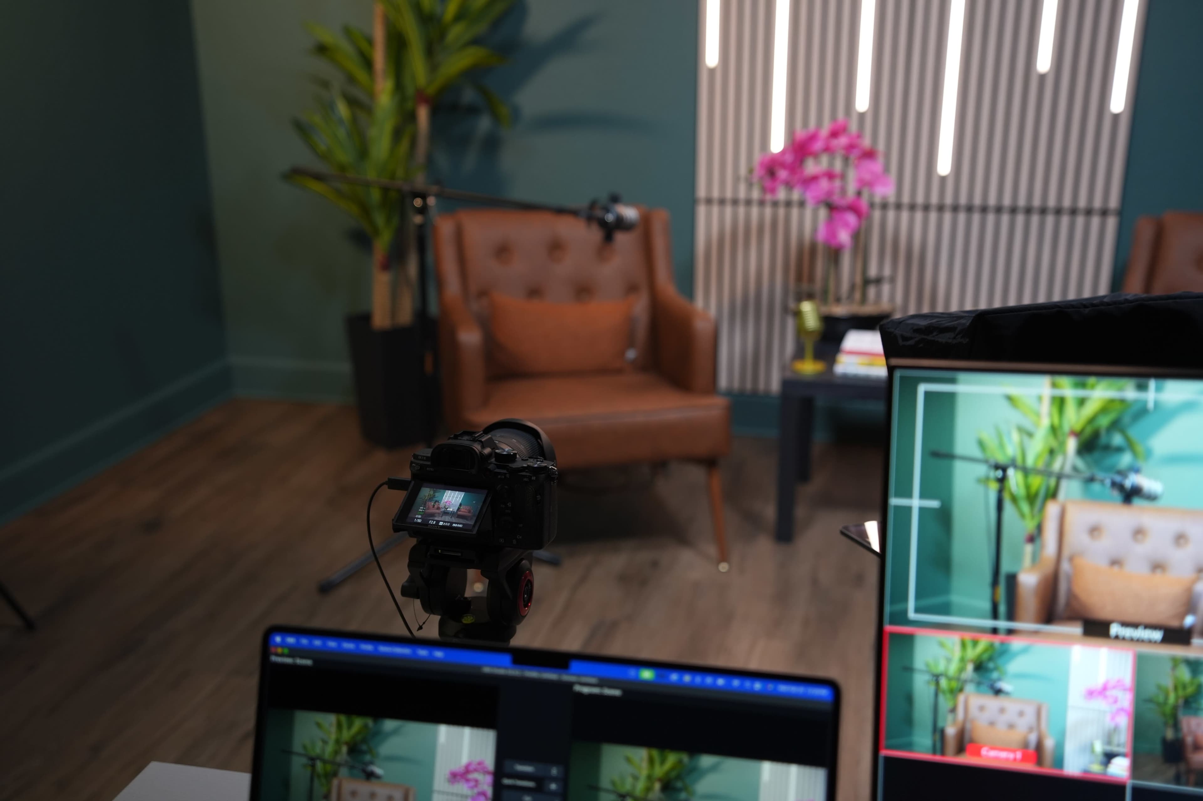 The image shows a recording setup featuring a brown upholstered chair, a camera on a tripod, and a computer monitor displaying a preview of the scene, alongside a potted plant and a table with books.