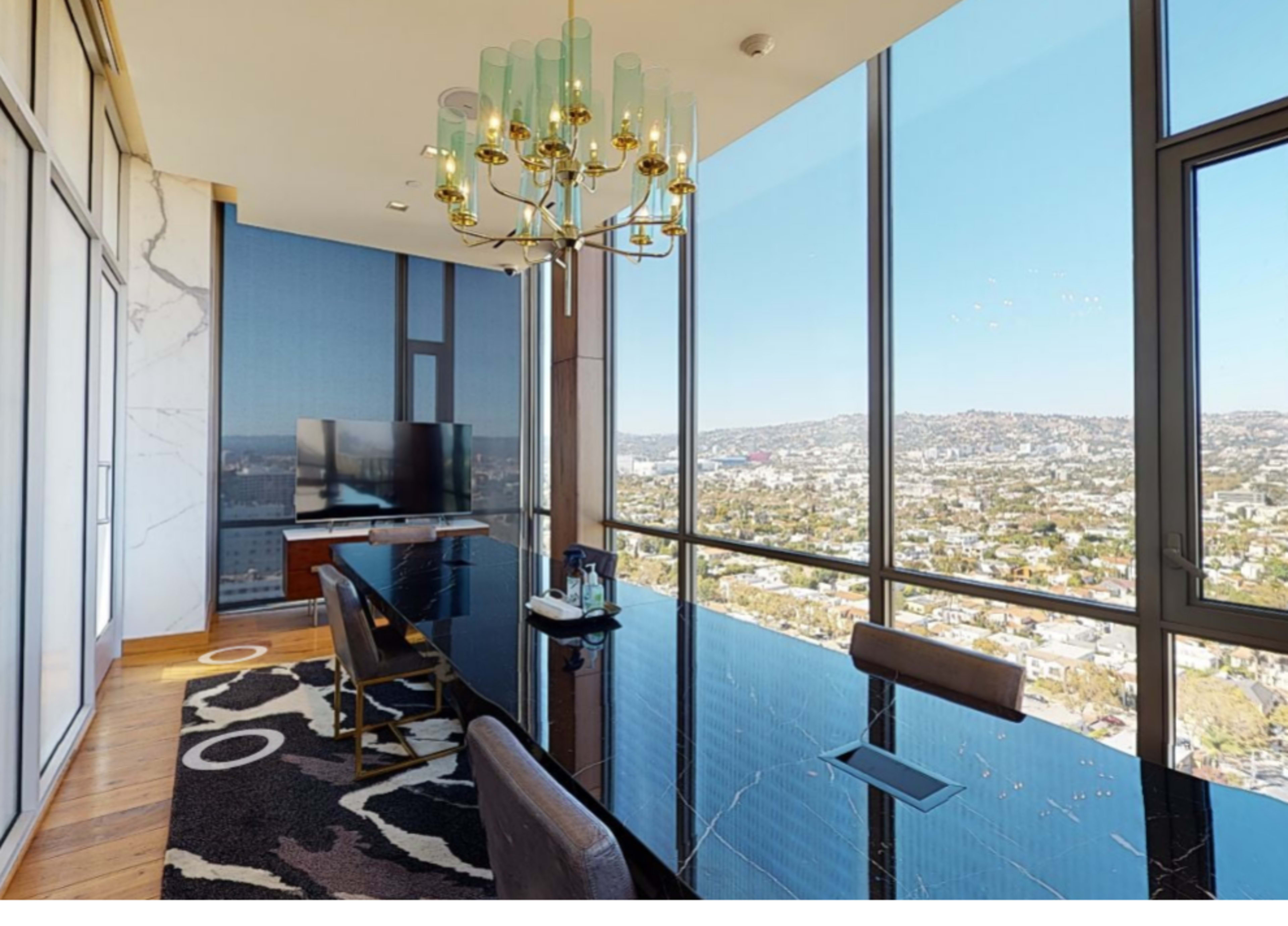 A modern conference room features a long black table, a chandelier, large windows with a city view, and a television on a wall.