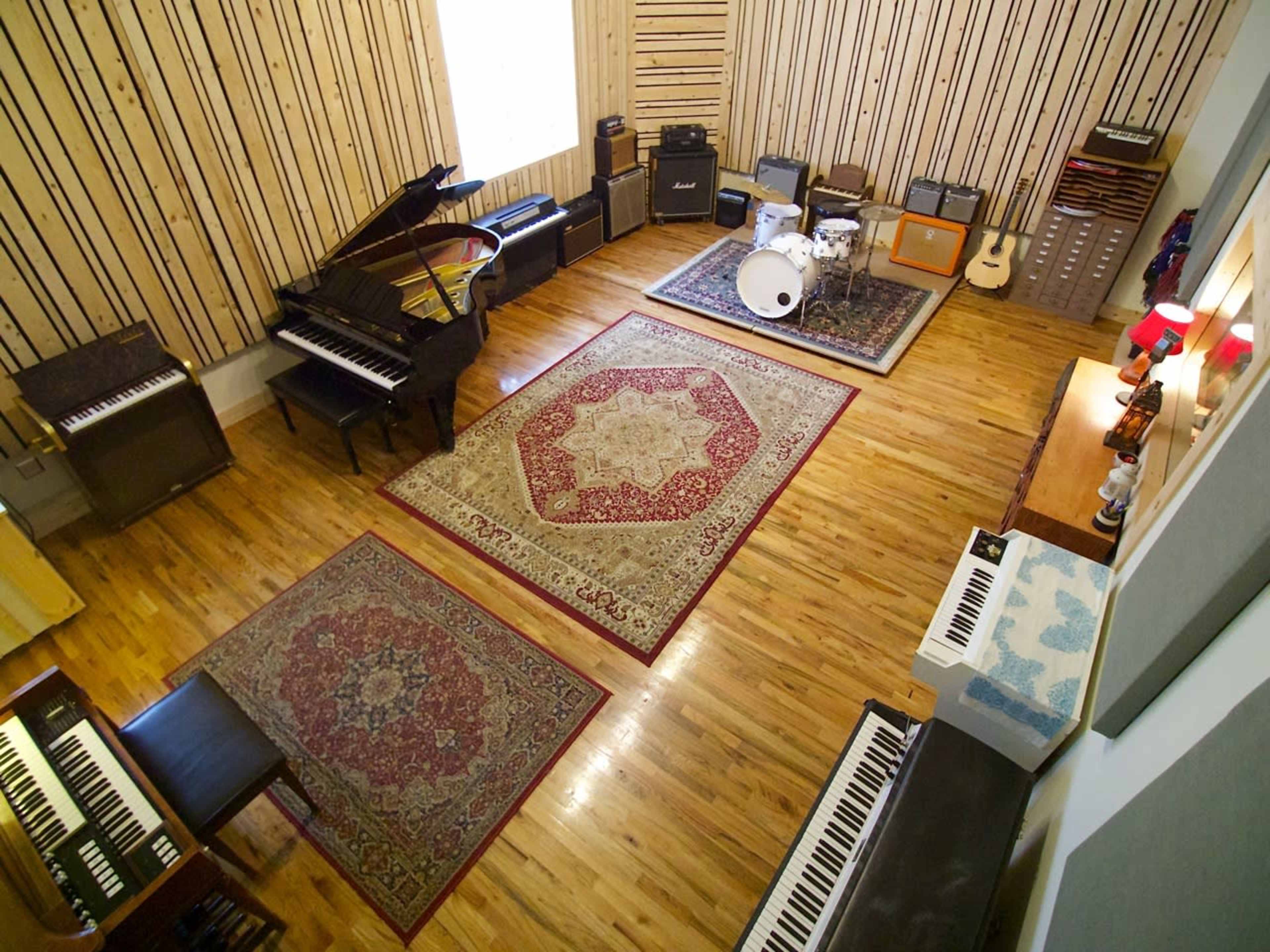 A spacious music studio featuring a grand piano, various amplifiers, a drum set, and several keyboards arranged on a hardwood floor with decorative rugs.