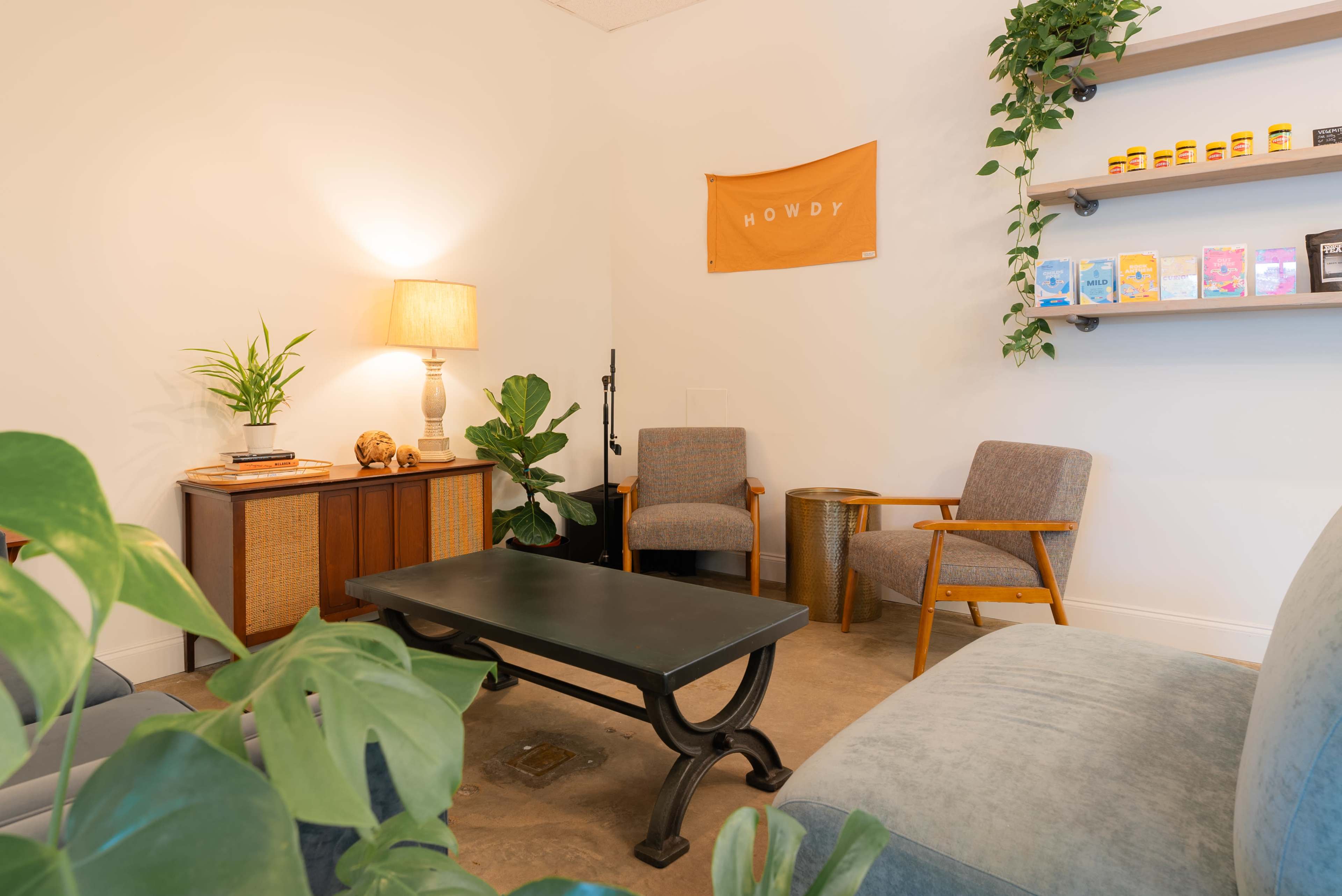 A cozy waiting area features two armchairs, a coffee table, a lamp, greenery, and shelves displaying various items and a wall hanging that says "HOWDY."