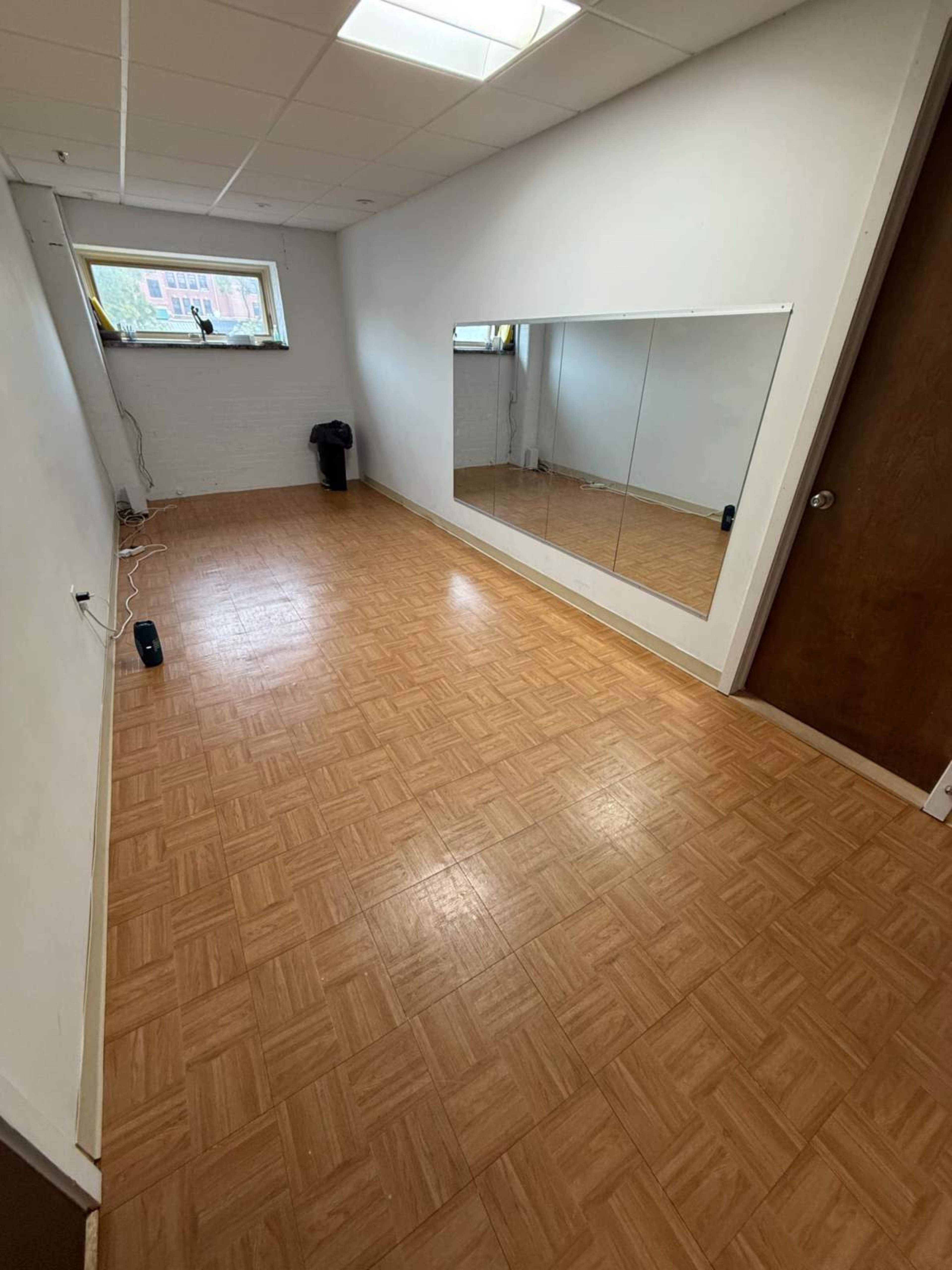 The image shows a vacant room with a wooden floor, a large mirror on one wall, and a window providing natural light.