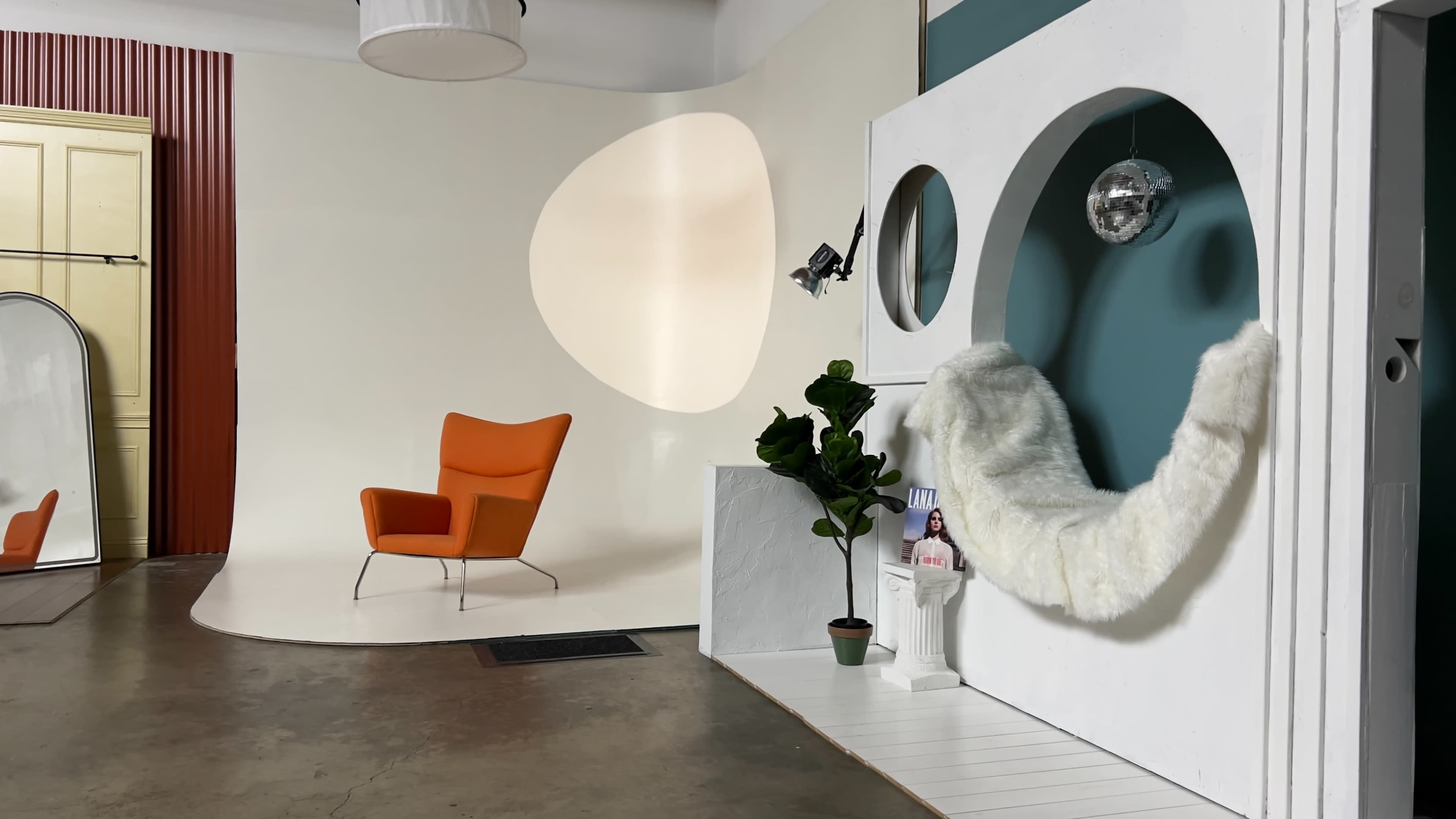 The image shows a modern interior space featuring a bright orange chair, a white wall with circular cutouts, and decorative elements like a potted plant and a disco ball.
