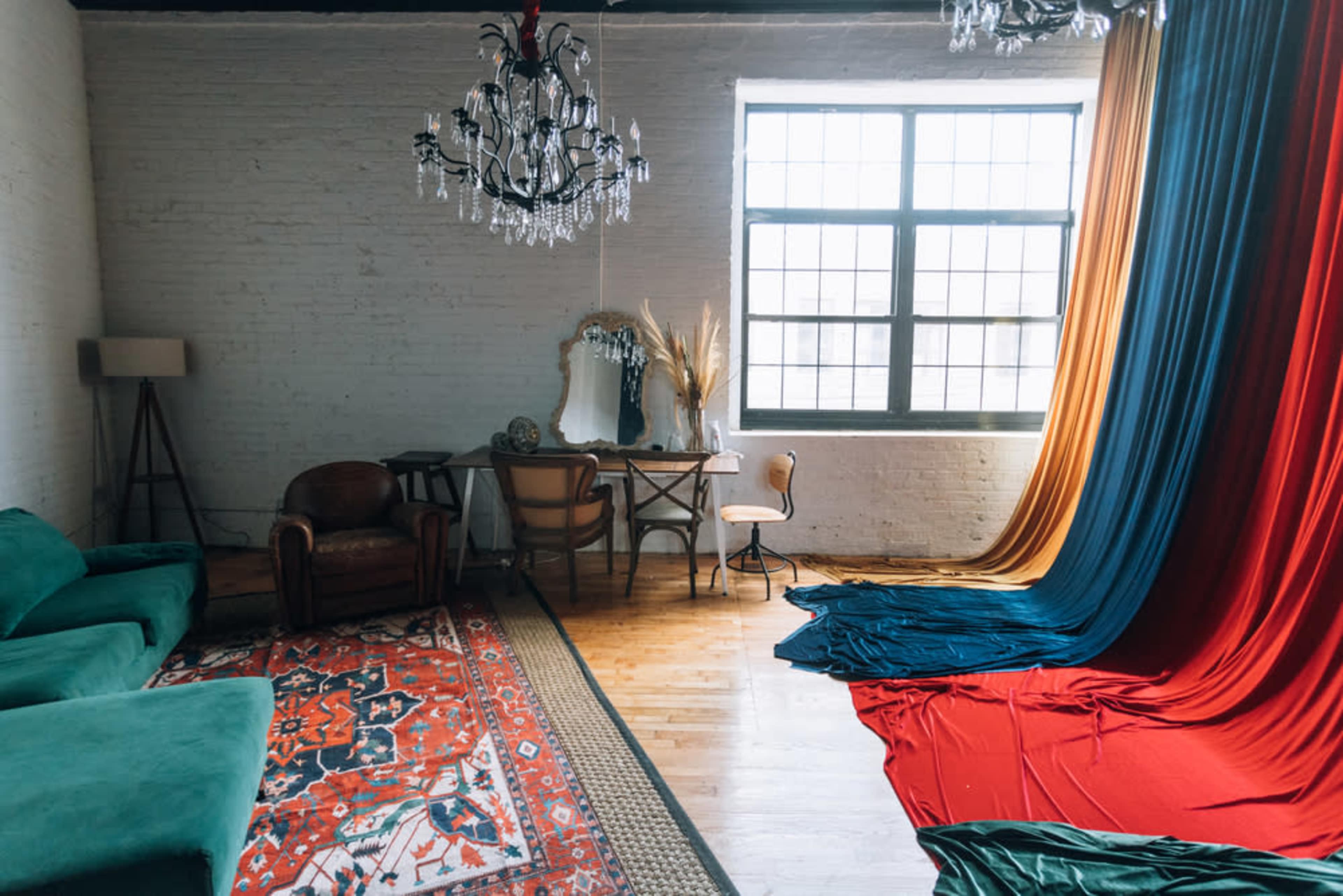 The image shows a spacious room featuring a mix of colorful drapes, a vintage mirror, a wooden table with a chair, and a green couch set against a backdrop of large windows.