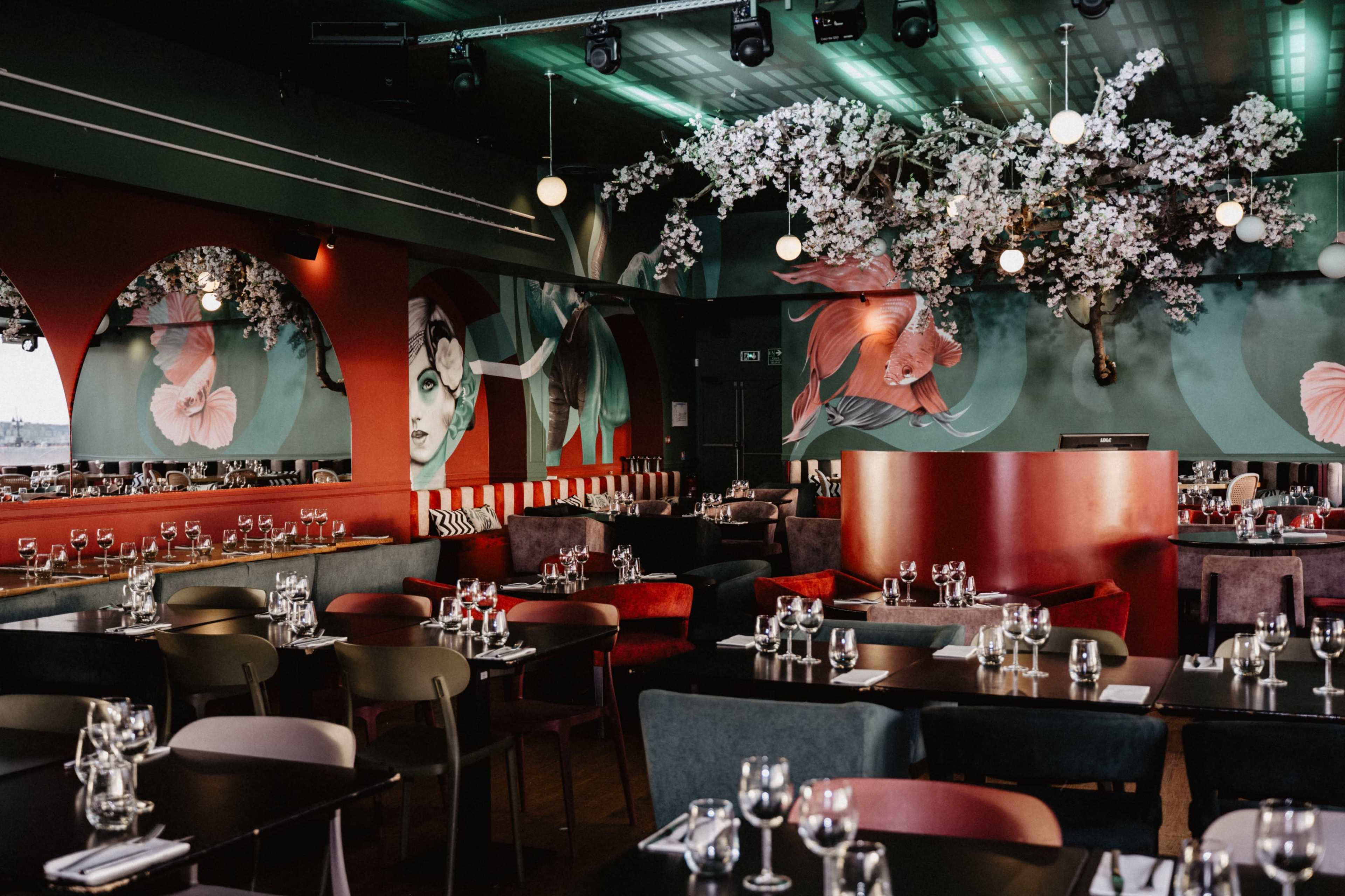 The image shows a modern restaurant interior featuring colorful decor, artistic wall murals, and tables set with glassware.