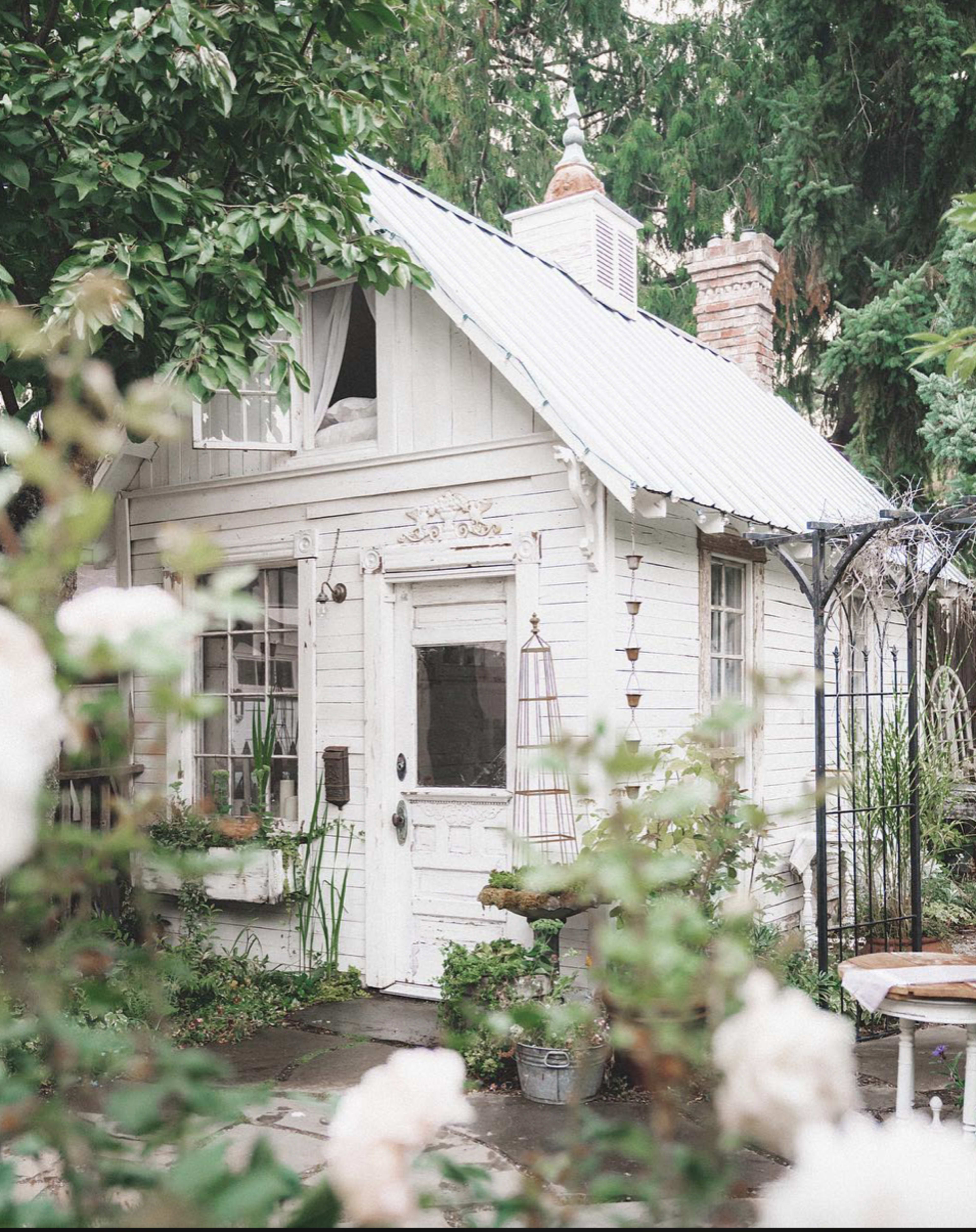 A small white cottage with a metal roof is surrounded by greenery and flowers in a serene outdoor setting.