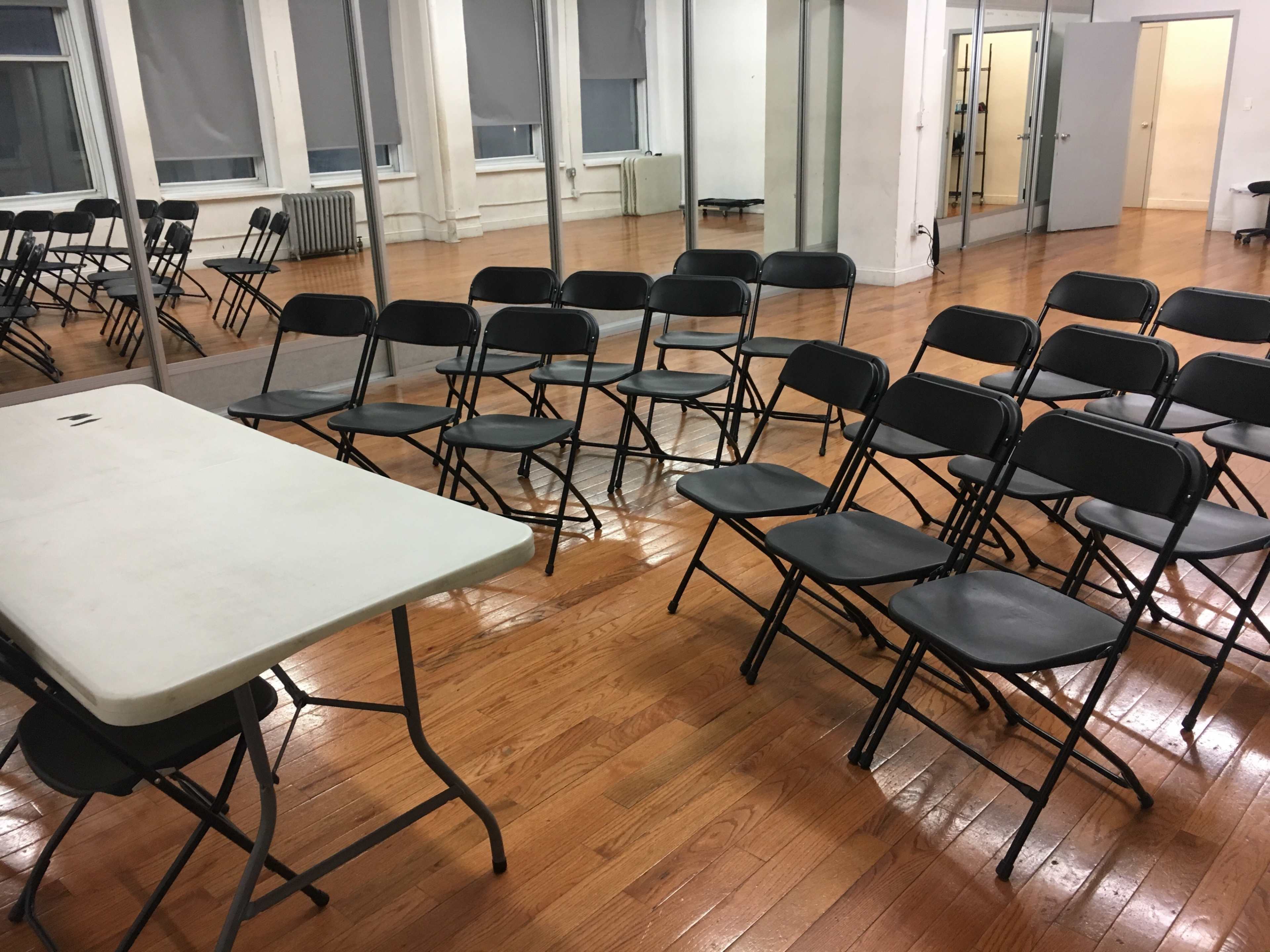 The image shows a room with wooden floors, several rows of black folding chairs, and a table set off to the side, all reflected in a wall of mirrors.