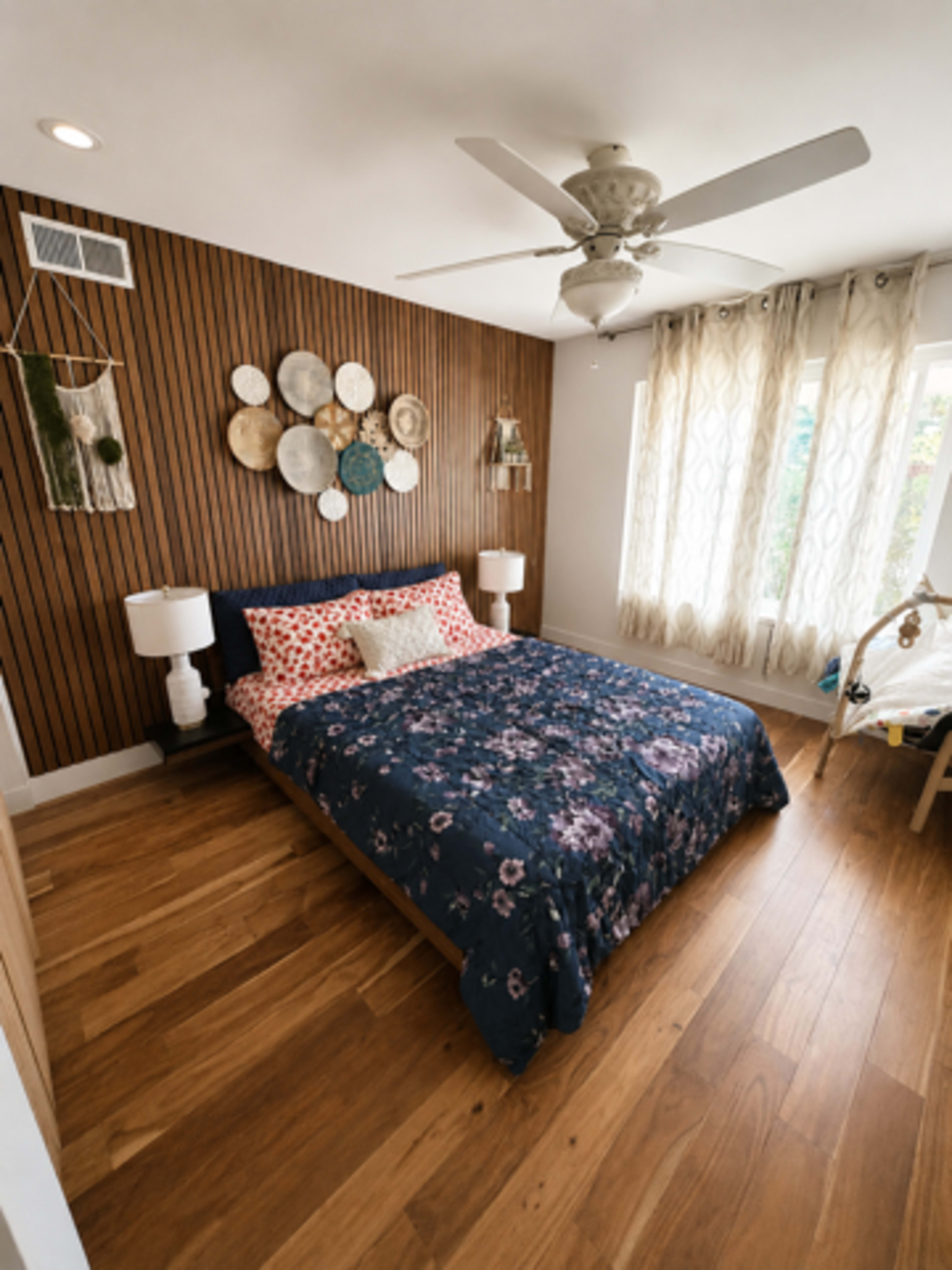 The image shows a cozy bedroom with a bed covered in a floral-patterned quilt, wooden walls, and decorative plates arranged above the bed.