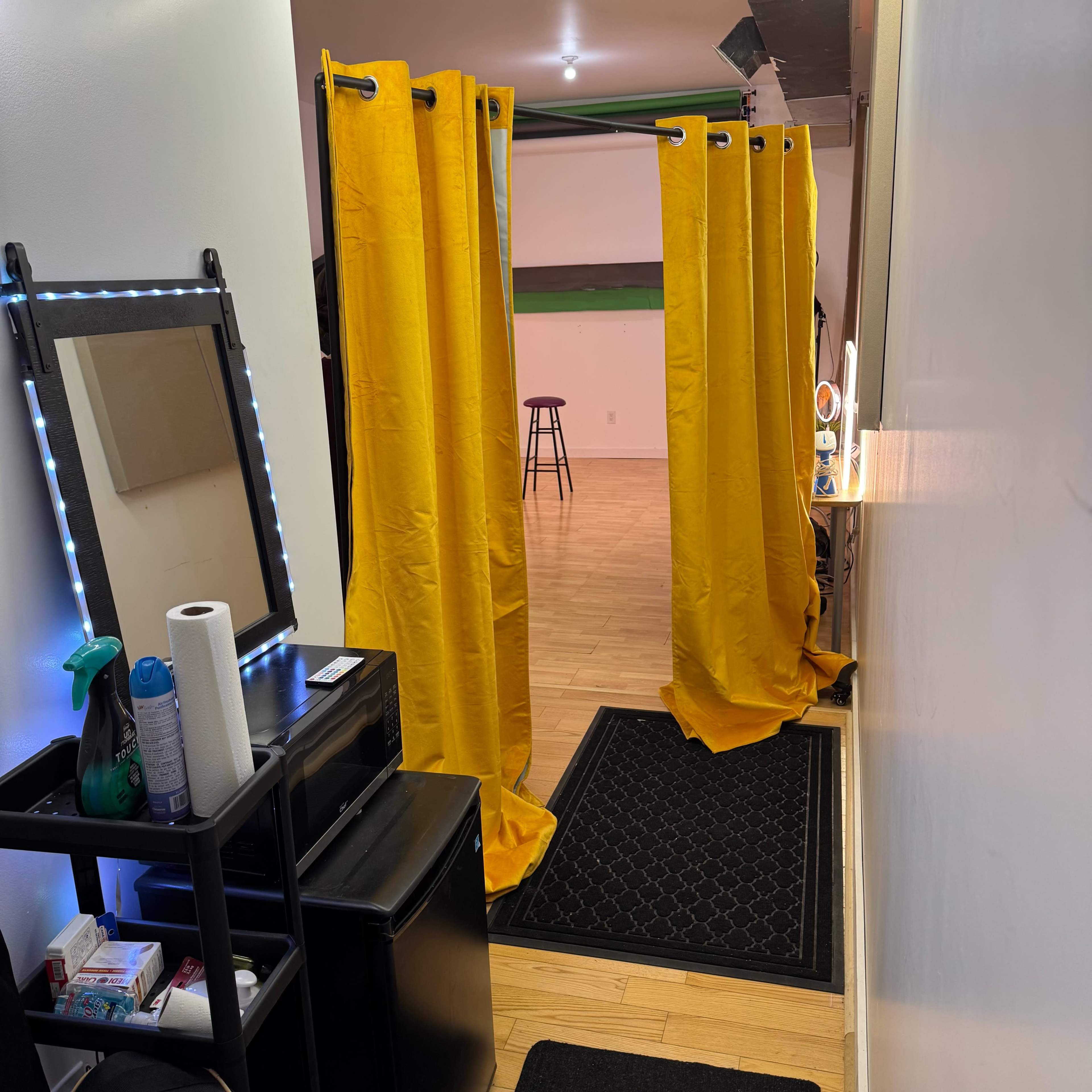 The image shows a narrow hallway with yellow curtains partitioning off an area, along with a mirror, a small table, and a mat at the entrance.