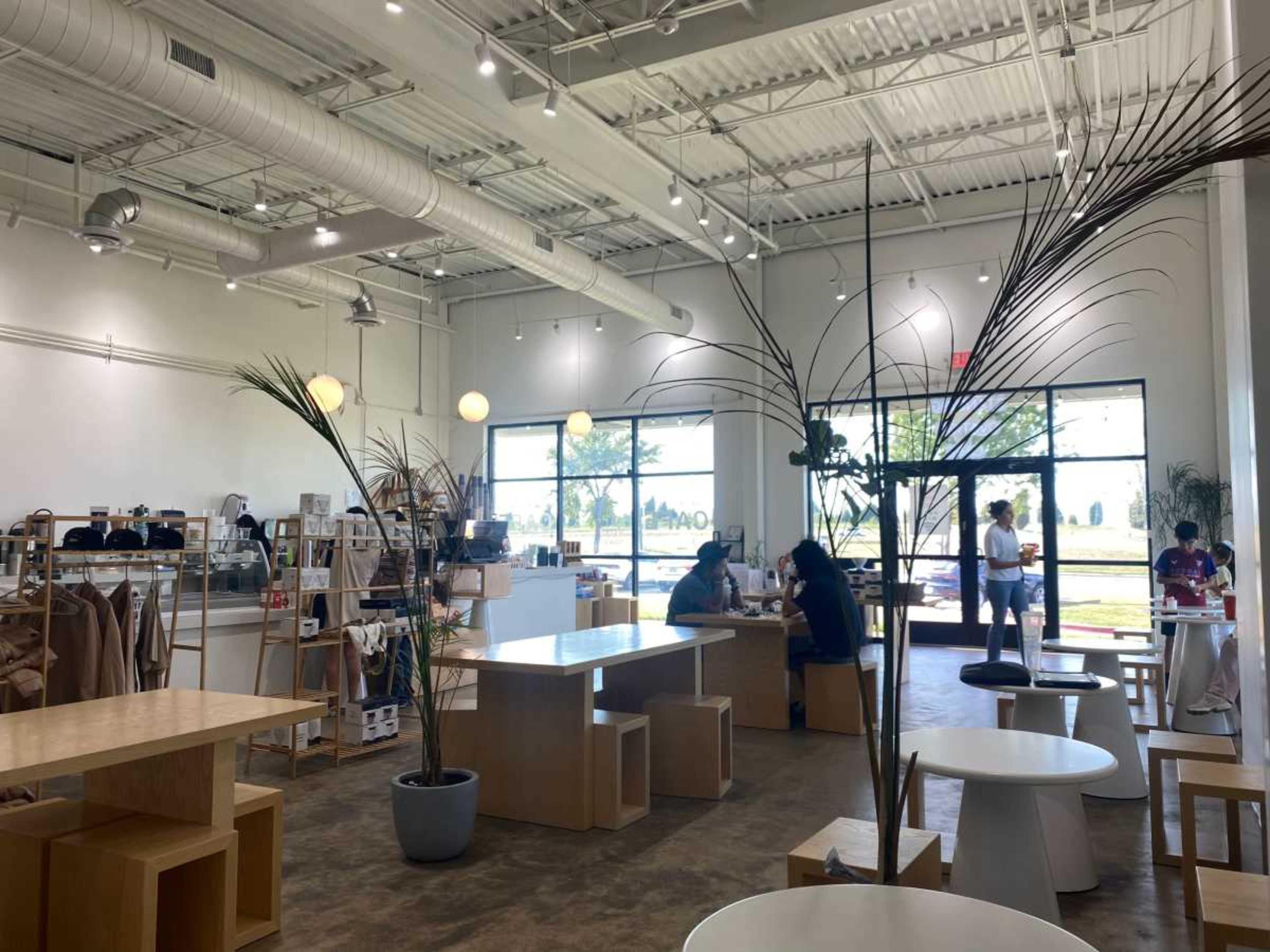 The image shows a modern café interior with several wooden tables and chairs, large windows letting in natural light, and patrons seated at various tables.