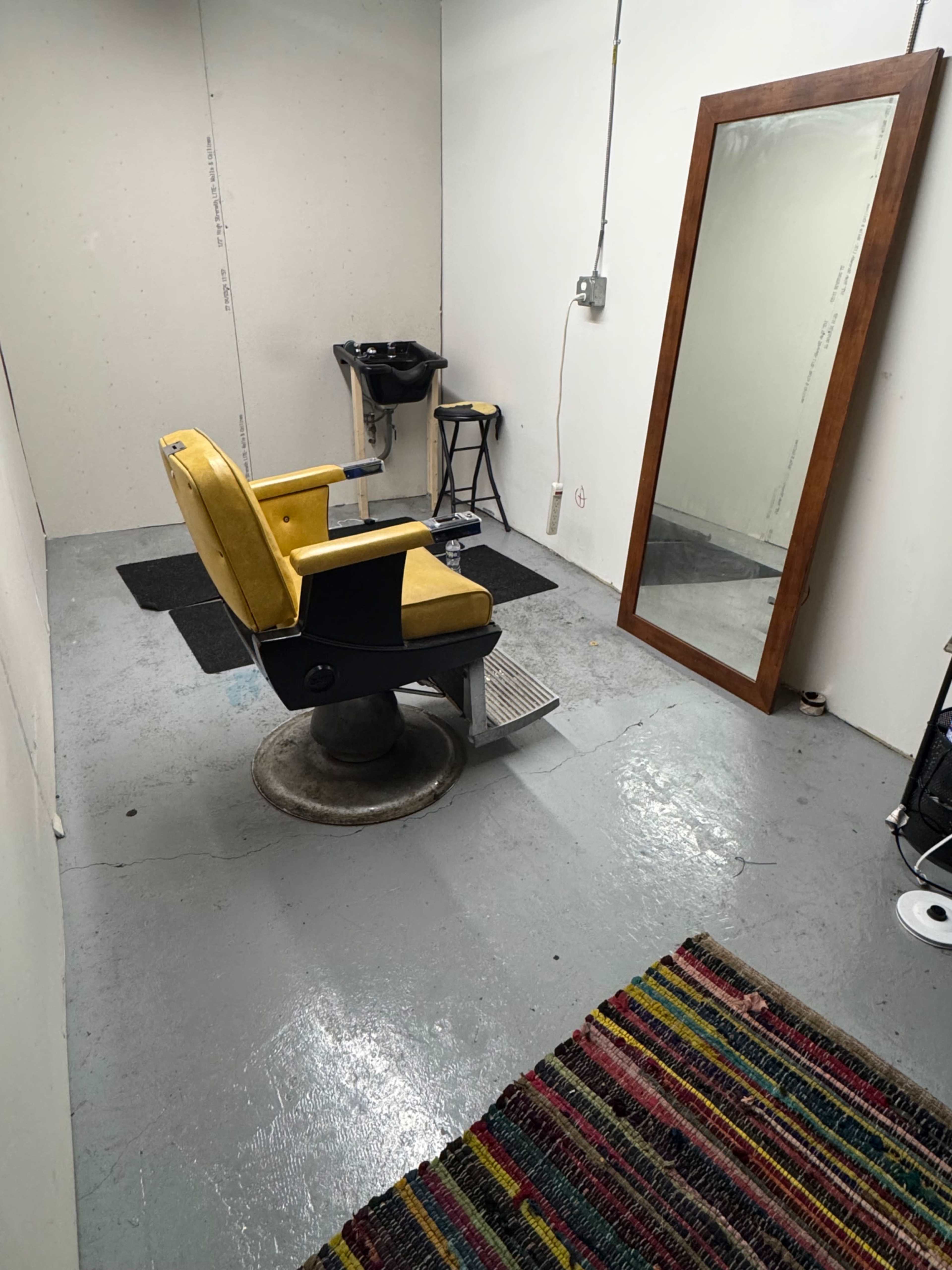 The image shows a small, minimally furnished room with a yellow barber chair, a mirror on the wall, and a shampoo basin in the corner.