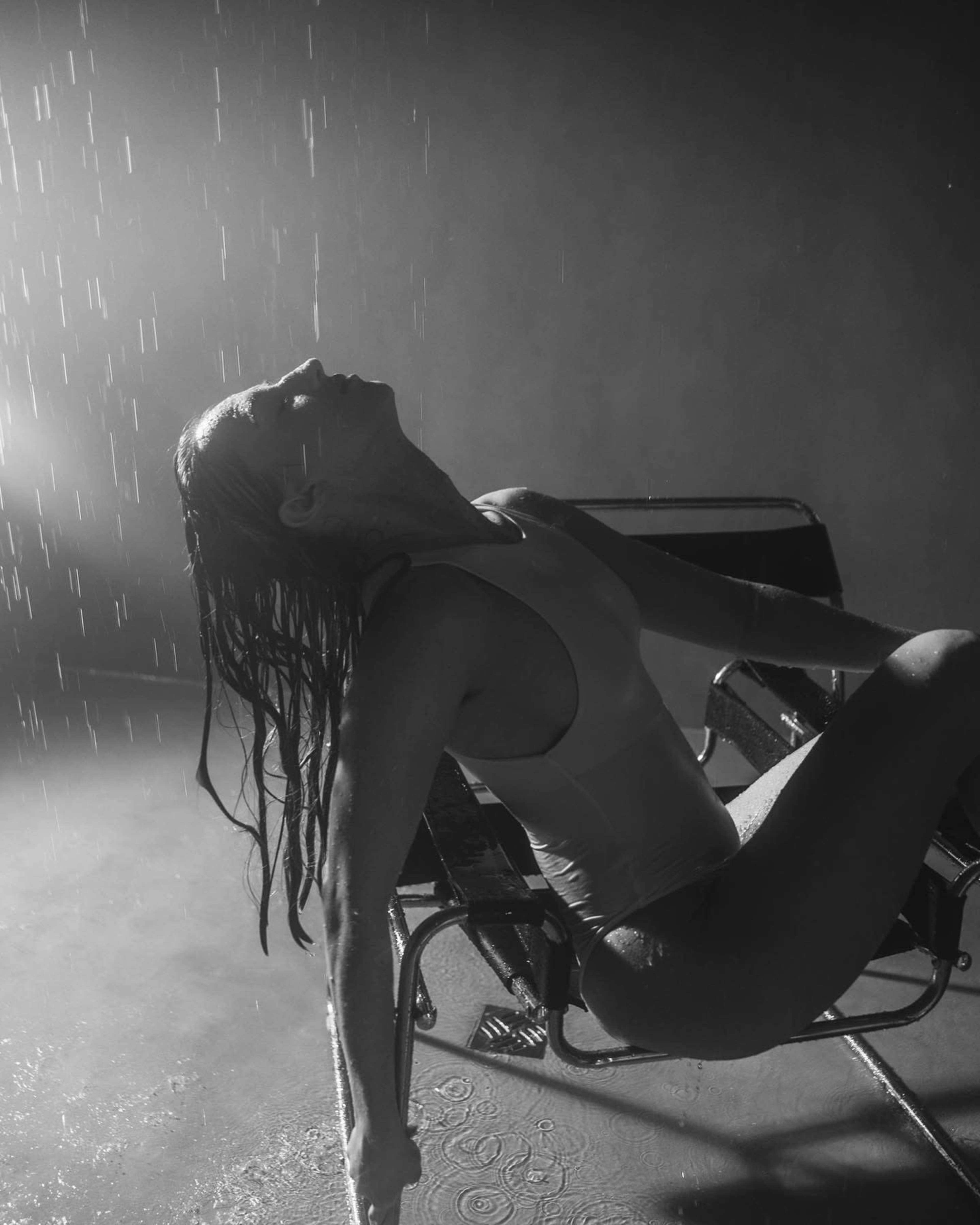 A woman with wet hair is sitting in a chair under falling water in a dimly lit space.